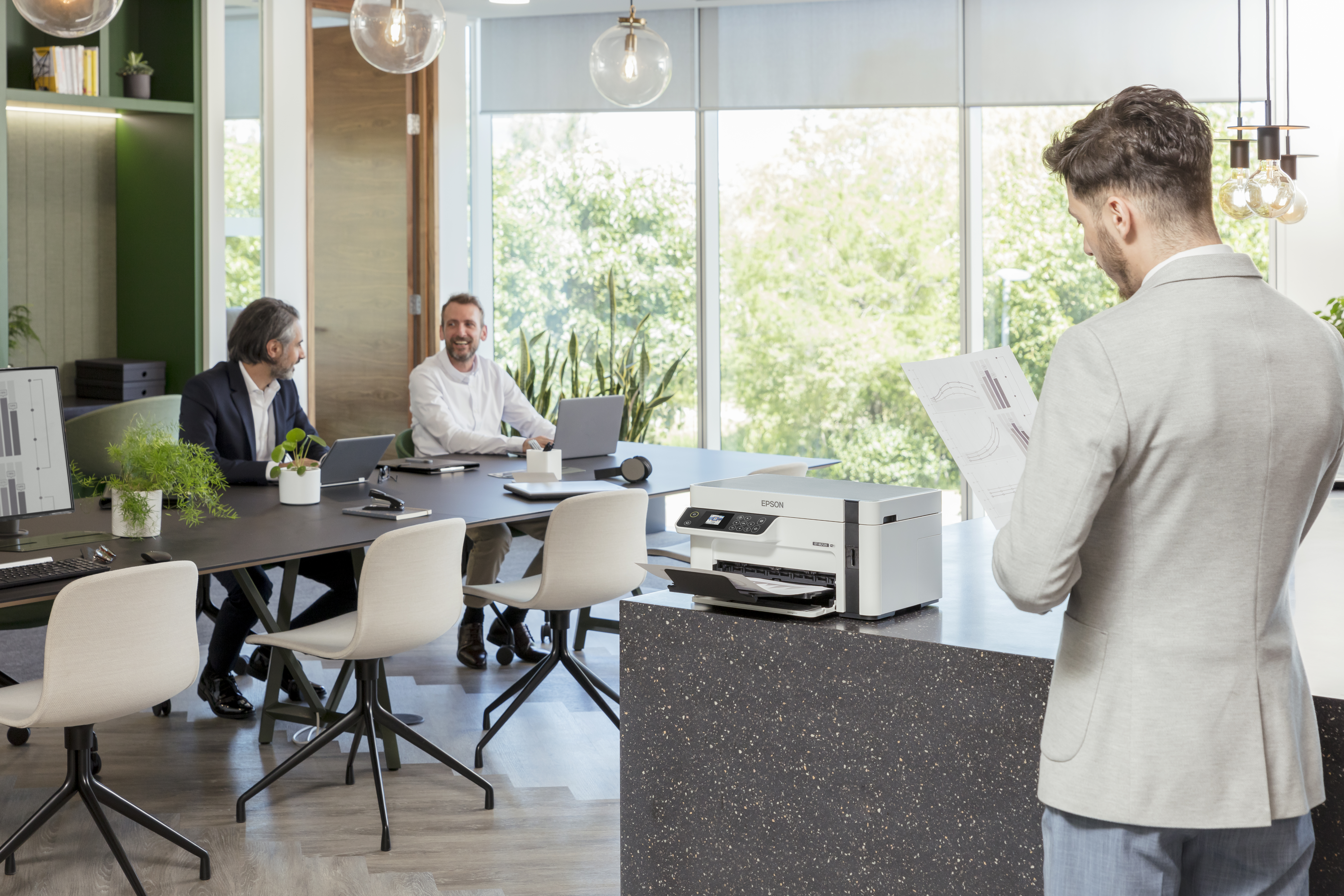 Geschäftstreffen in modernem Büro: 2 Männer am Tisch, 1 Mann prüft Unterlagen.