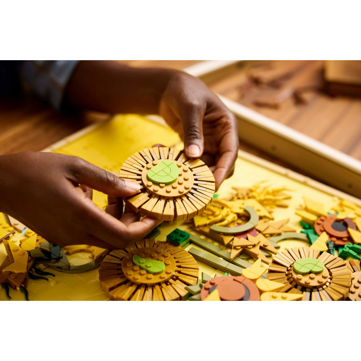 Mani che assemblano un girasole con mattoncini lego marroni e verdi su base gialla.