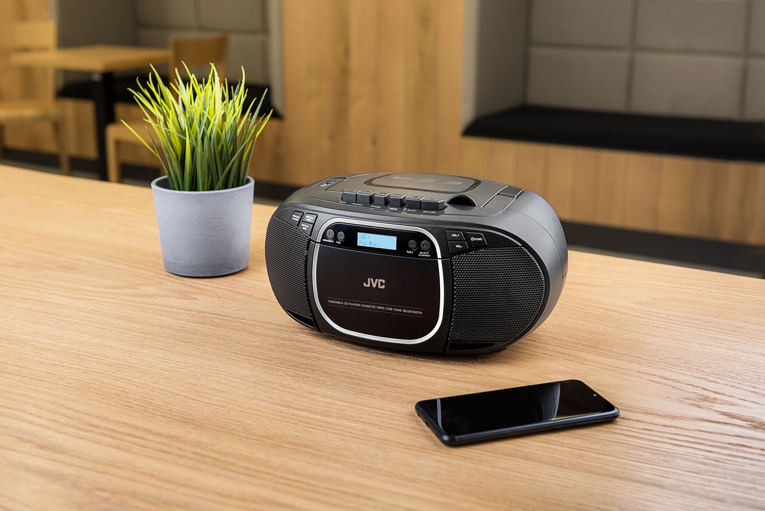 Egy fekete JVC boombox CD-lejátszóval faasztalon áll egy zöld növényt és egy okostelefont.