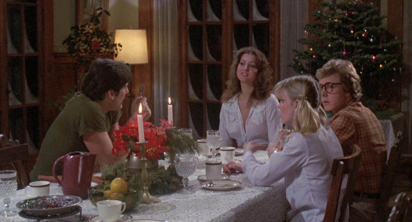 Vier Personen sitzen an einem festlich gedeckten Tisch mit Kerzen und einem Weihnachtsbaum im Hintergrund.