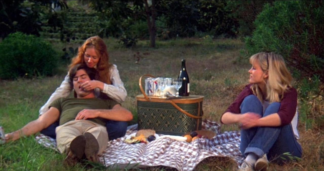 Drei Leute machen ein Picknick im Freien. Ein Mann liegt, eine Frau füttert ihn, eine andere schaut zu.