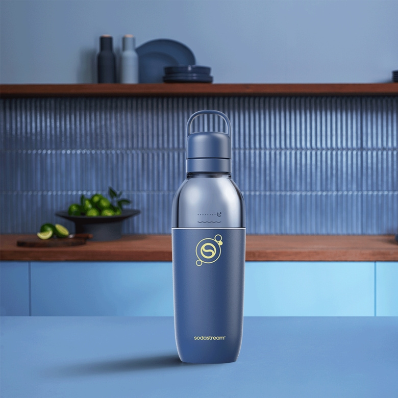 Niebieska butelka Sodastream stoi na niebieskim stole przed kuchenną półką z limonkami.