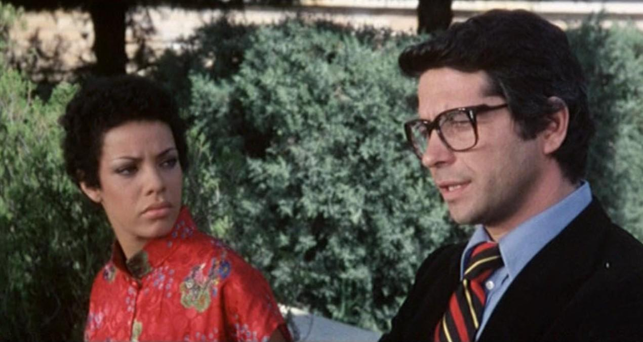 Eine Frau mit kurzen Haaren und ein Mann mit Brille sitzen vor einem Busch.