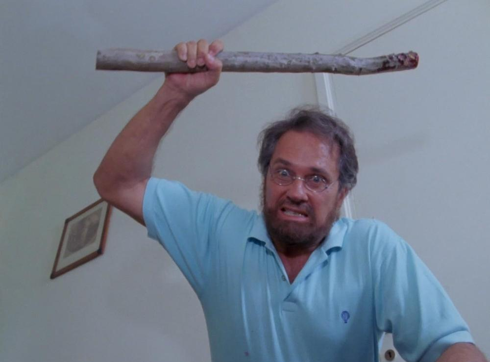 Mann mit Holzstock hebt ihn über dem Kopf, Grimasse schneidend.