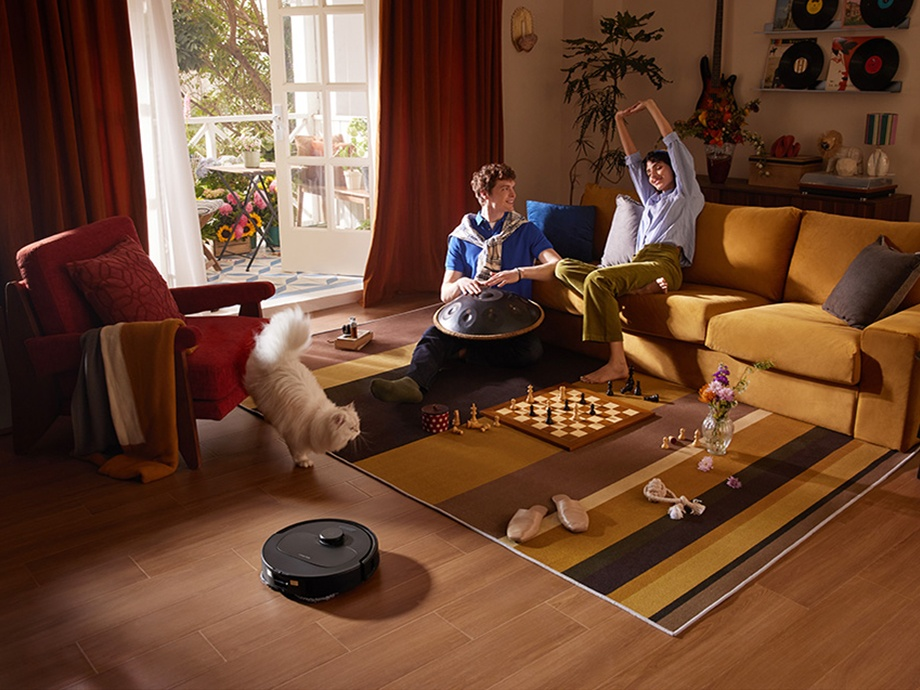 Dwie osoby bawią się w salonie, obecne są kot i robot sprzątający.