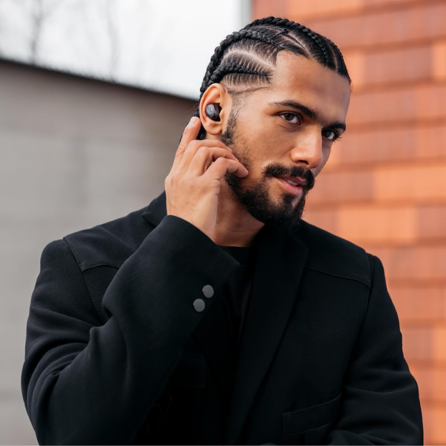 Uomo con capelli intrecciati e barba indossa cappotto nero e auricolari.