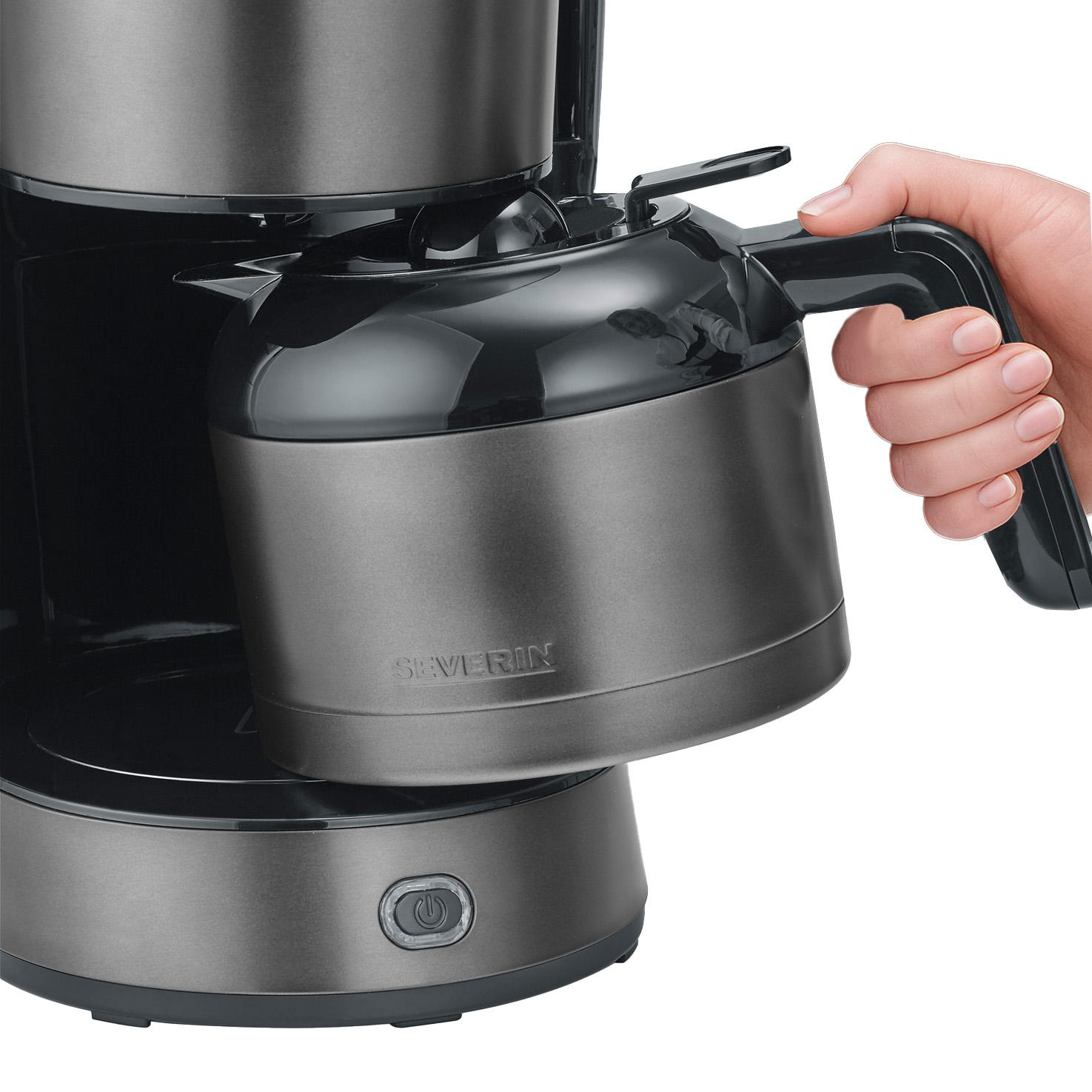 Eine Hand greift den schwarzen Griff einer grauen Kaffeekanne und setzt sie in eine Kaffeemaschine.