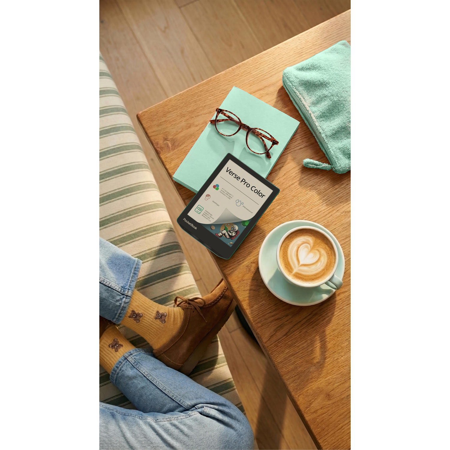 Een boek, bril en koffie liggen op een houten tafel naast de benen van een persoon.