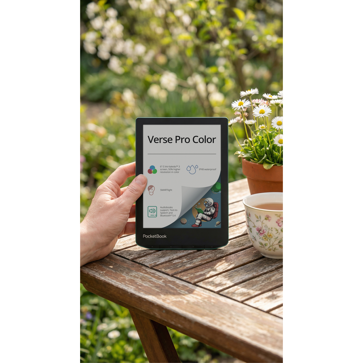 Een hand houdt een e-reader met kleur vast in de tuin. Een theekopje staat ernaast.