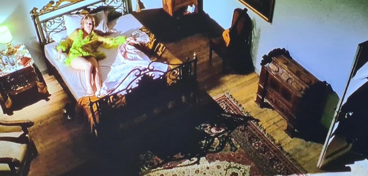 Eine Frau in einem grünen Feder-Kimono liegt auf einem Bett in einem opulenten Zimmer.