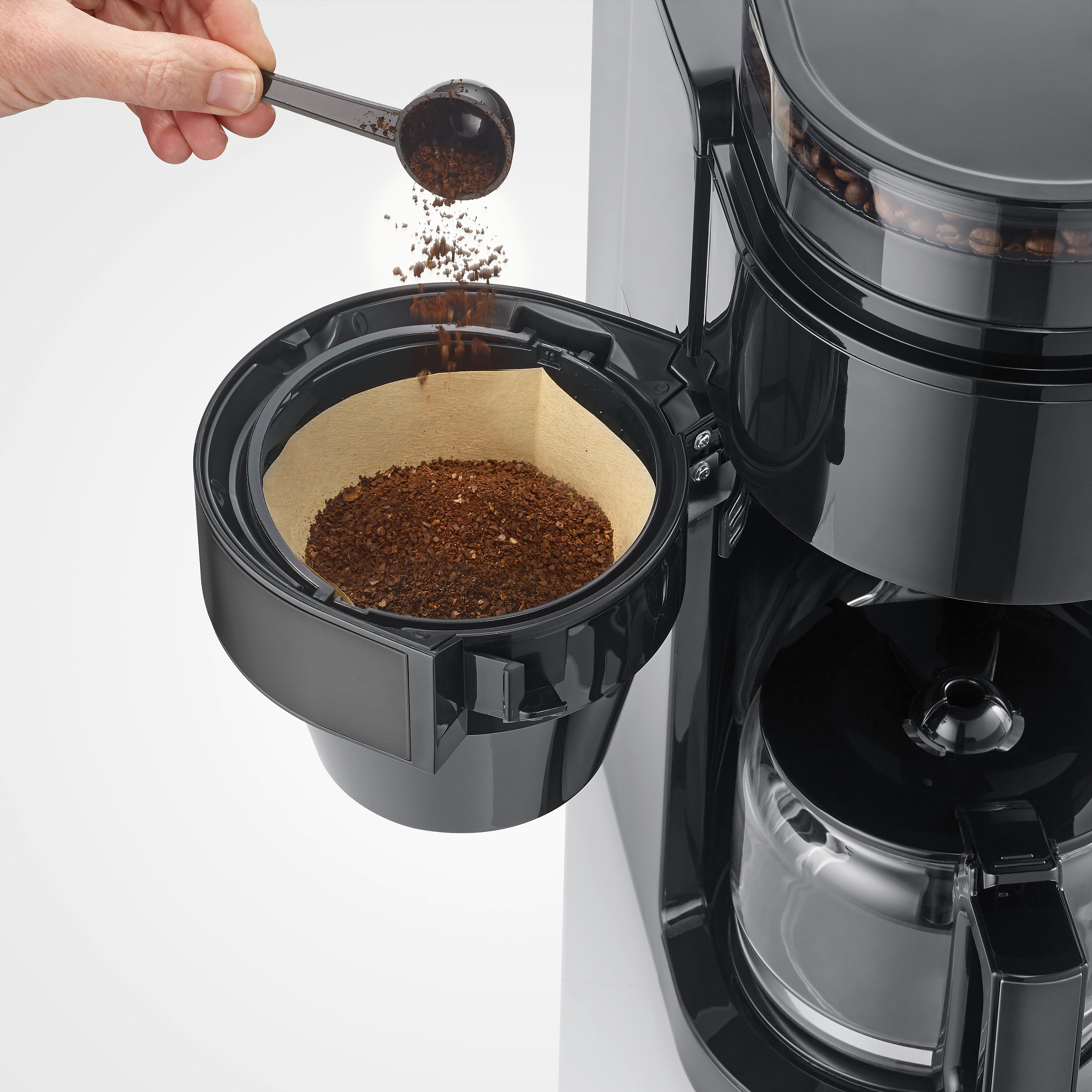 Eine Hand schaufelt Kaffeepulver in den Filterkorb einer schwarzen Kaffeemaschine.