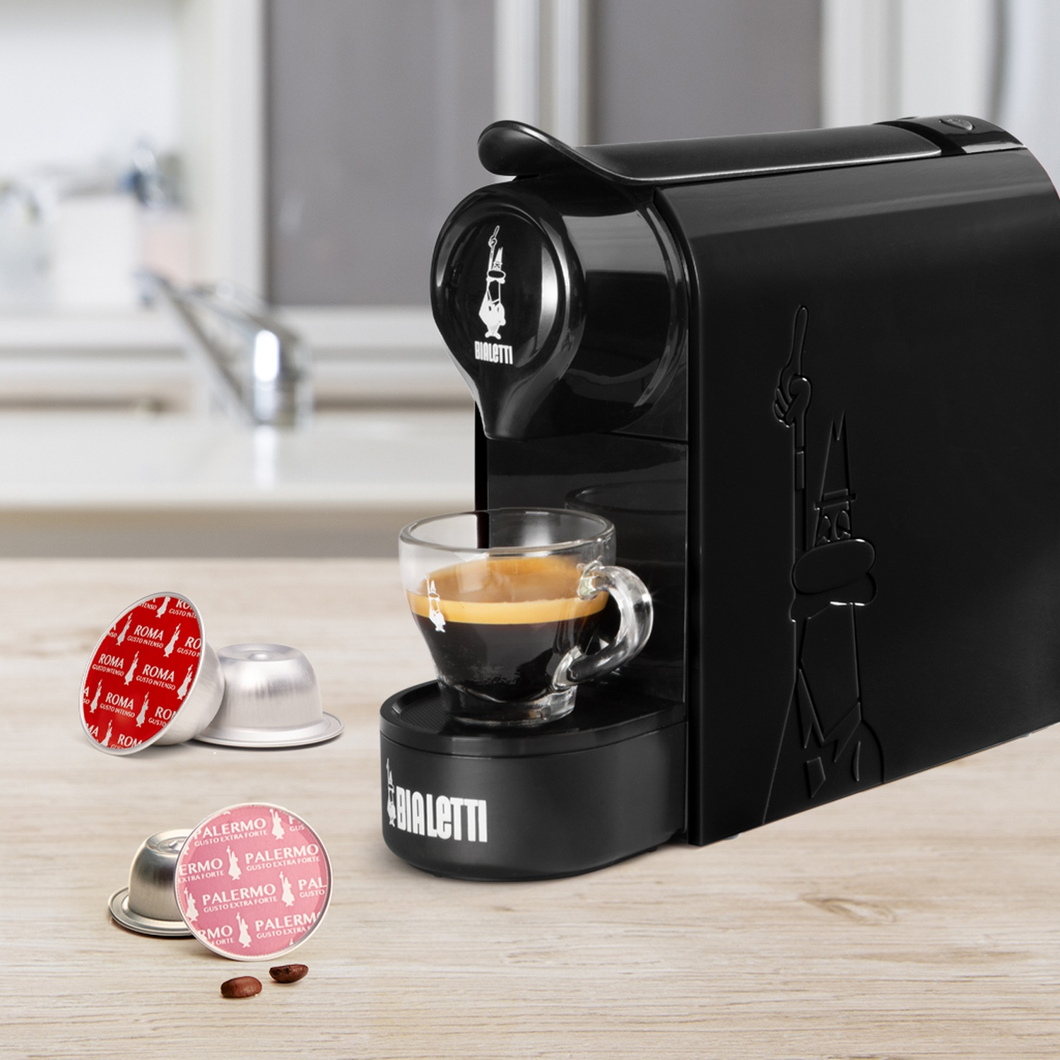 Macchina da caffè Bialetti nera con tazza di espresso e capsule di caffè su tavolo in legno.