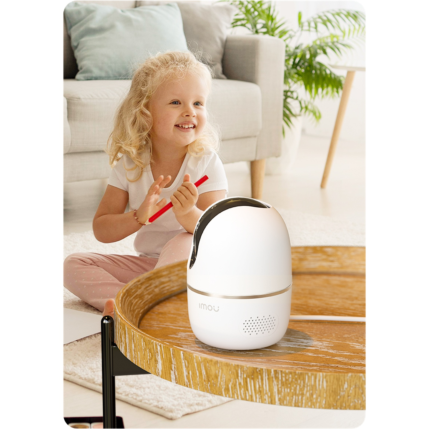 Une petite fille assise par terre tient un crayon rouge. Un babyphone est sur une table en bois.
