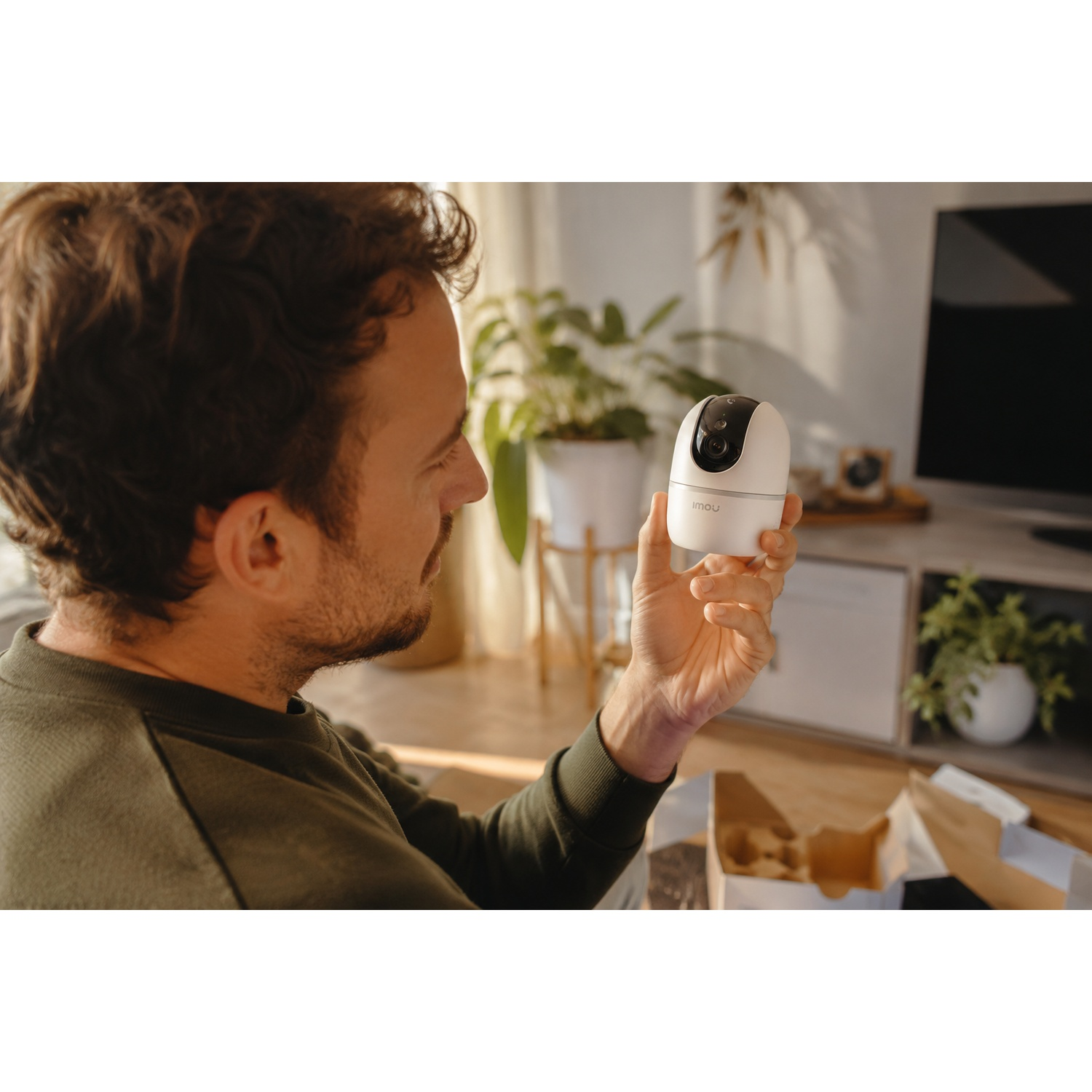 Homme tenant une caméra de sécurité blanche, avec emballage et plantes en arrière-plan.