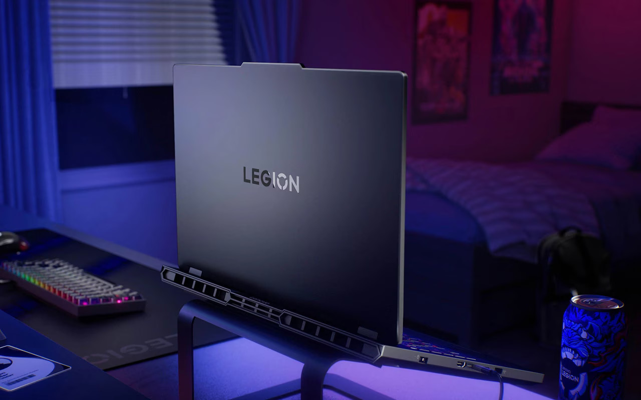 Laptop Lenovo Legion na biurku z klawiaturą i myszką. Łóżko w tle.