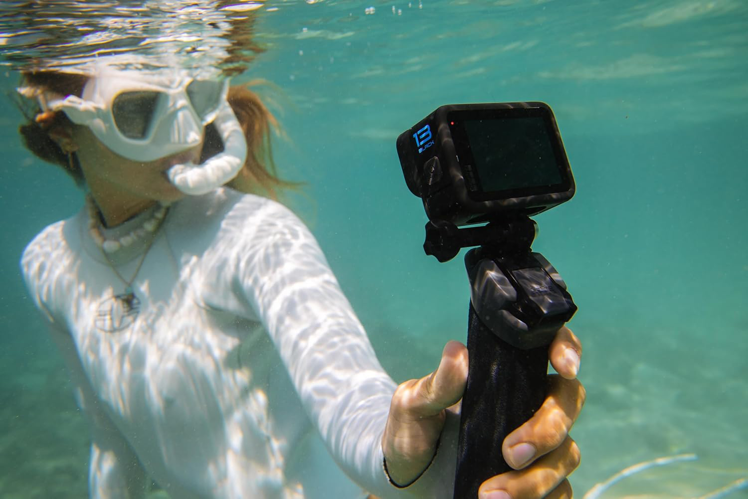 Neoprén ruhás, maszkos személy úszik víz alatt, GoPro kamerát tartva.