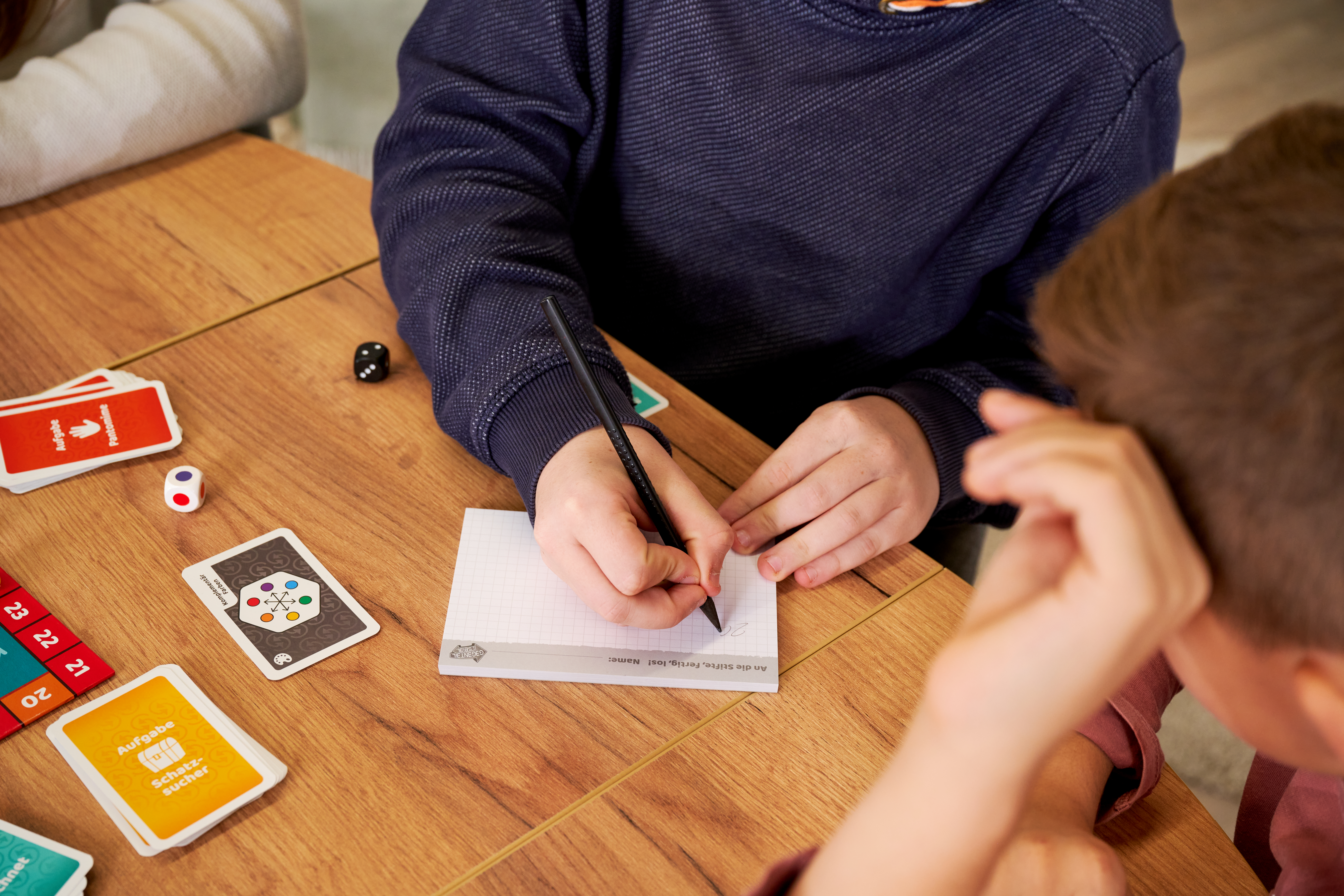 Kinder spielen ein Brettspiel mit Karten, Würfeln und Stiften auf einem Holztisch.