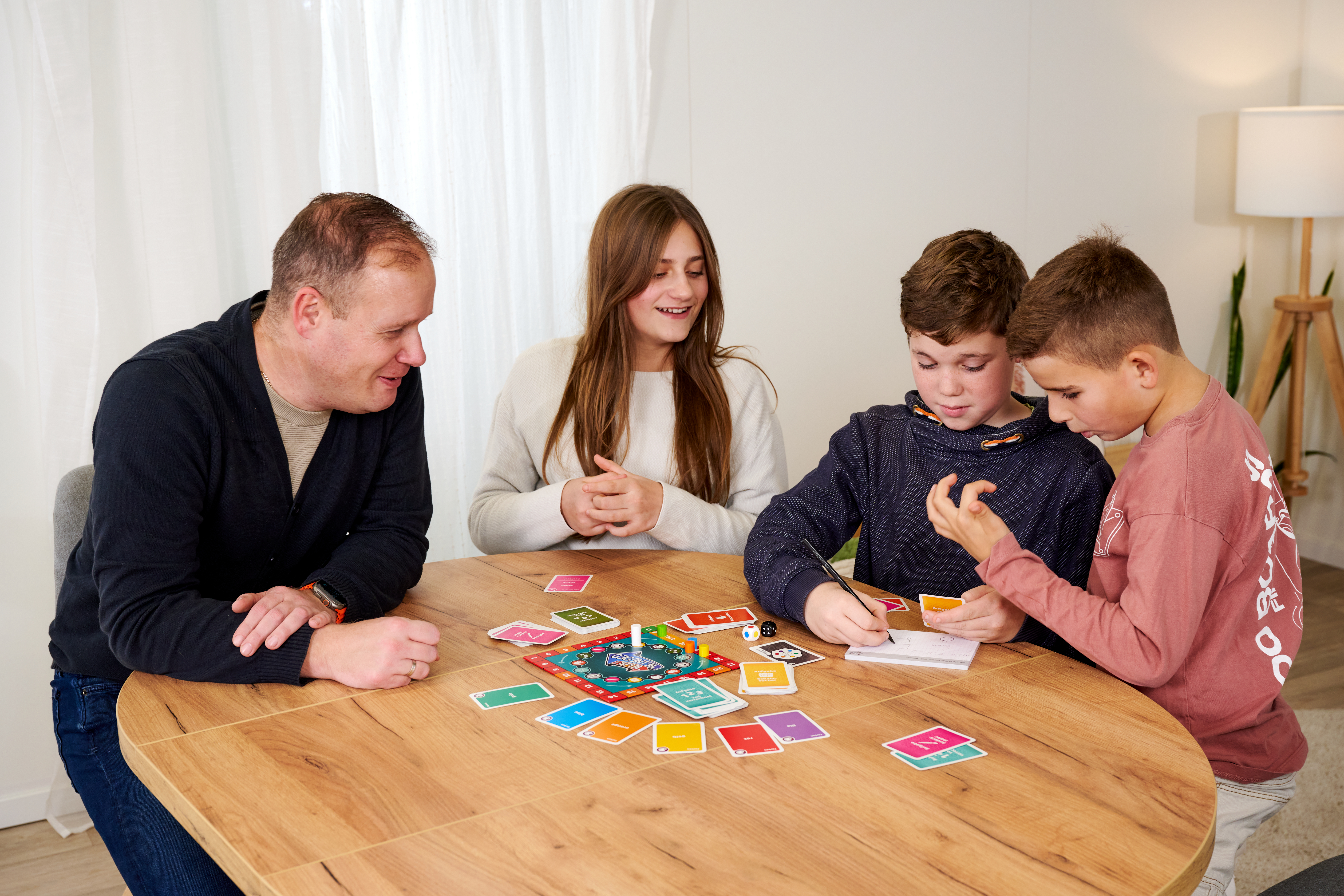 Familie spielt Brettspiel am runden Holztisch. Kinder schreiben und schauen auf Karten.
