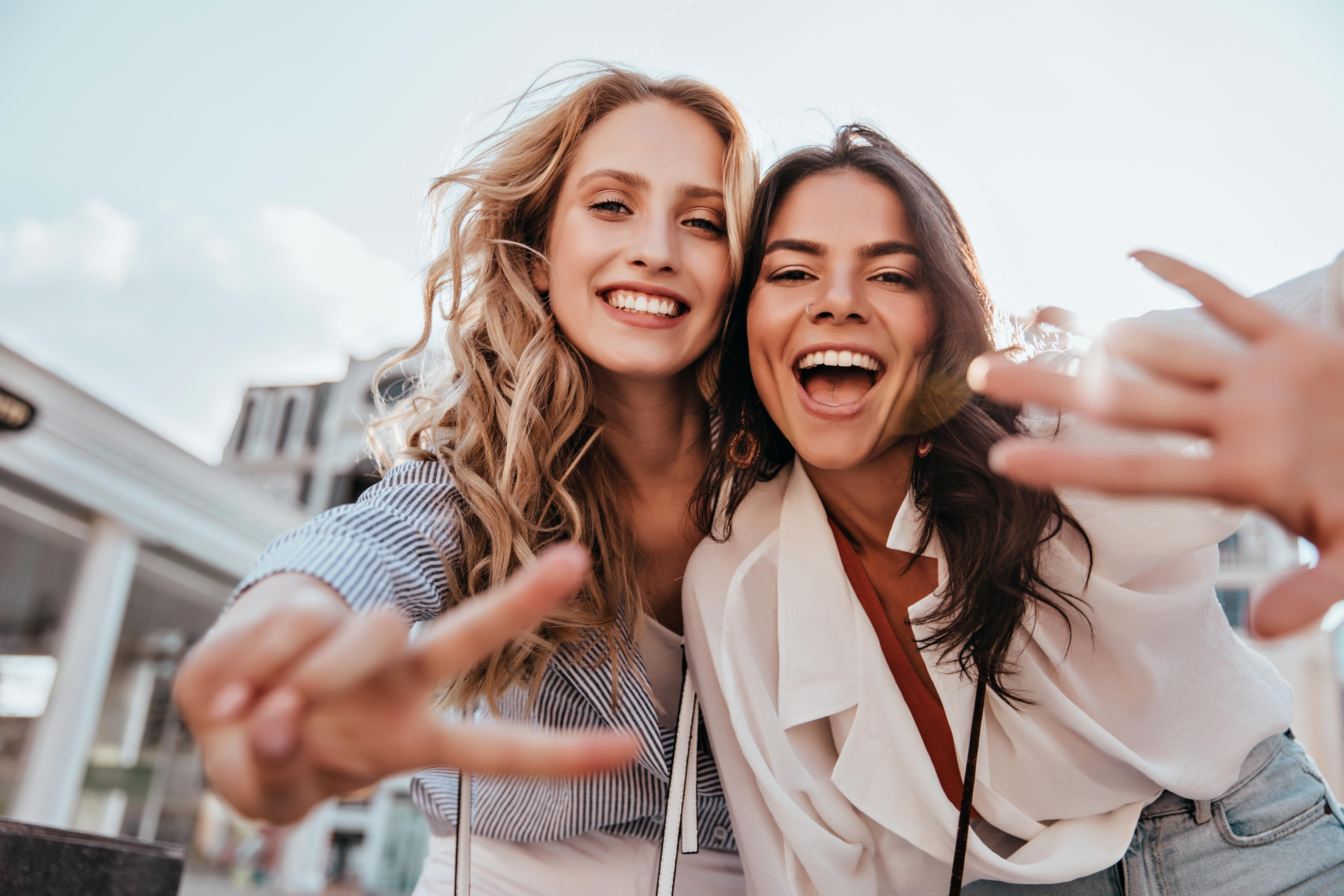 Zwei lachende Frauen machen ein Selfie vor einer unscharfen städtischen Kulisse.
