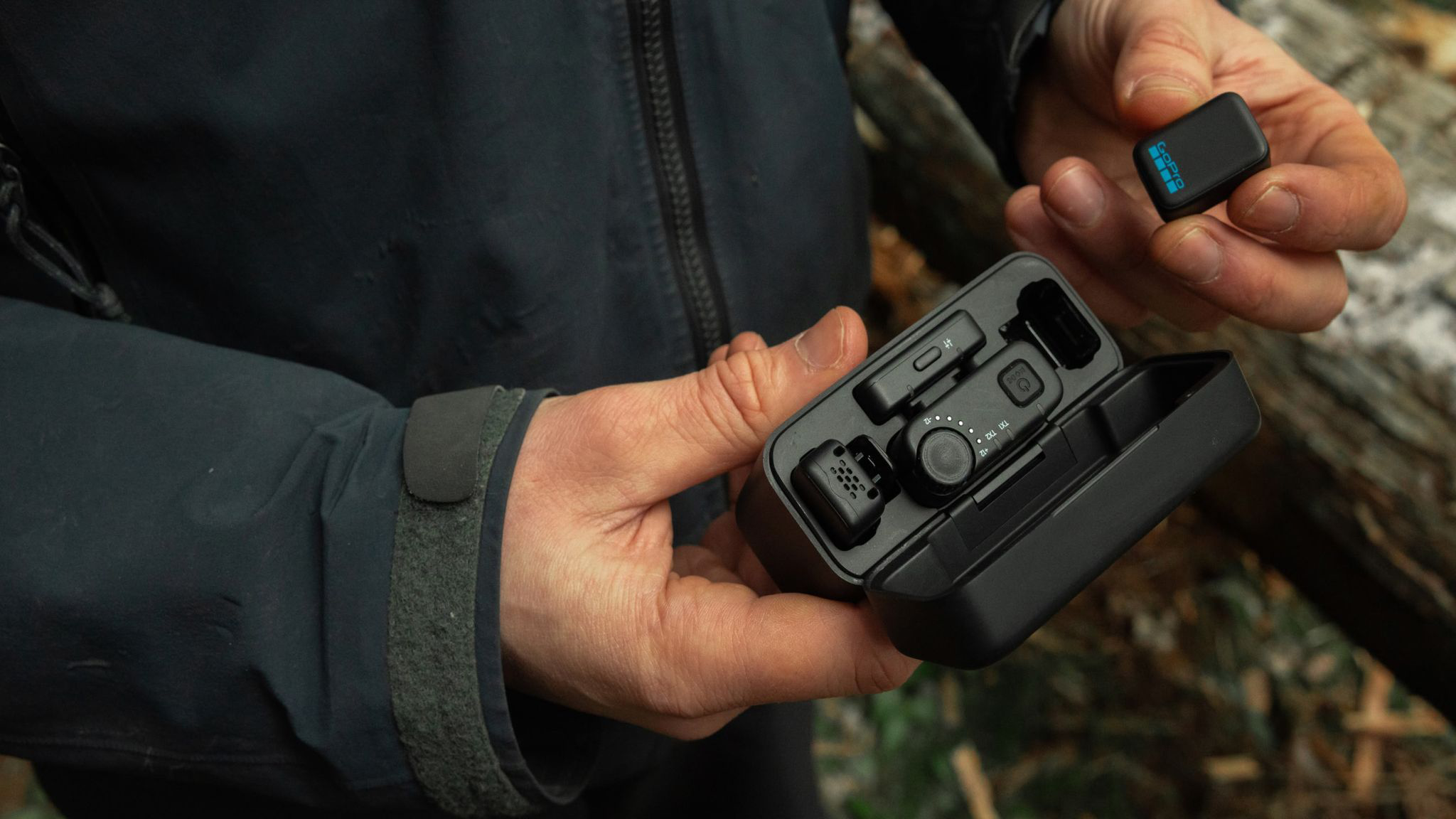 Egy kéz fekete GoPro mikrofont és töltőtokot tart tartozékokkal.