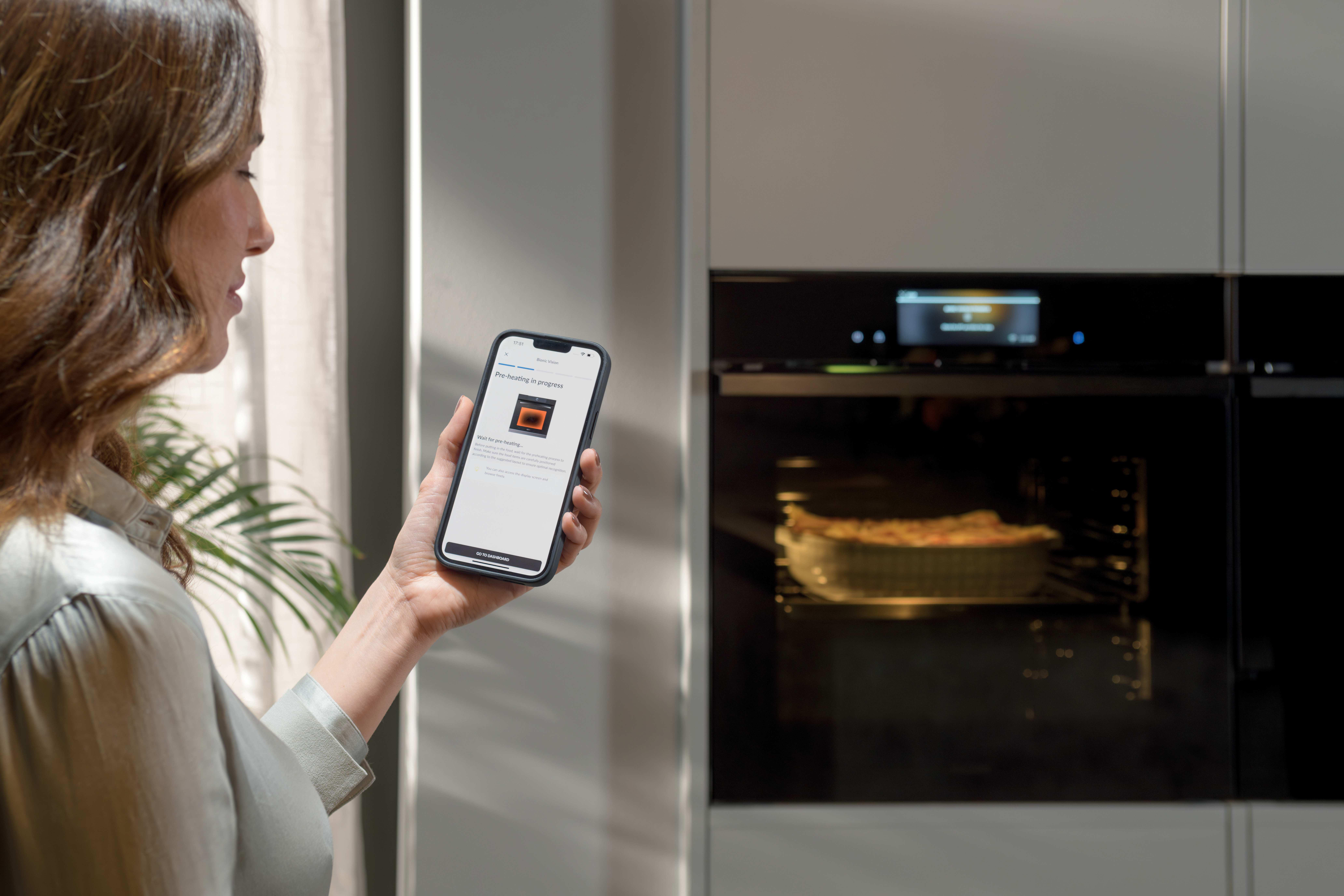 Eine Frau hält ein Smartphone mit einer Ofen-App, während ein Ofen vorheizt.