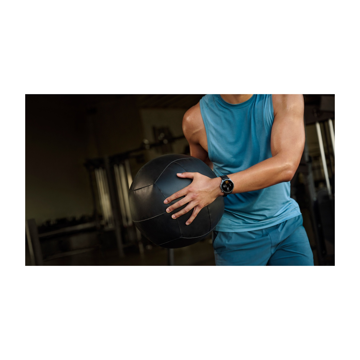 Uomo in palestra tiene palla medica nera, indossa canottiera blu e pantaloncini blu.