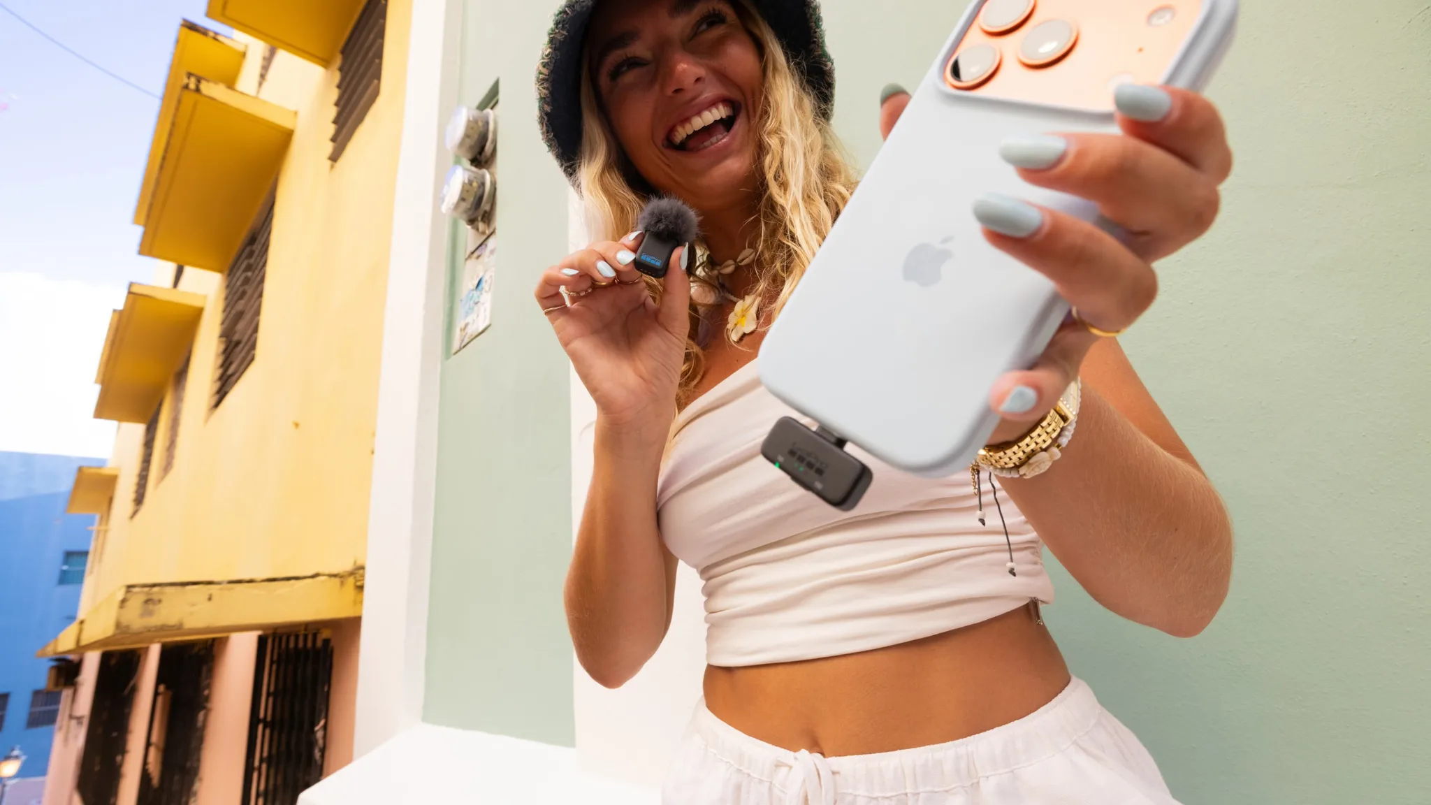 Egy mosolygó nő világoskék iPhone-t és mikrofont tart egy színes utcán.
