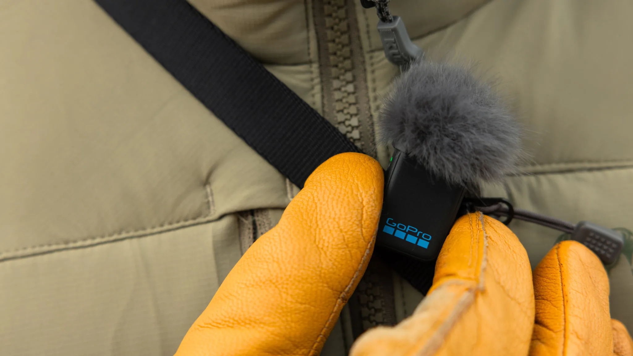 Sárga kesztyűs kezek GoPro mikrofont tartanak egy khaki dzsekin.