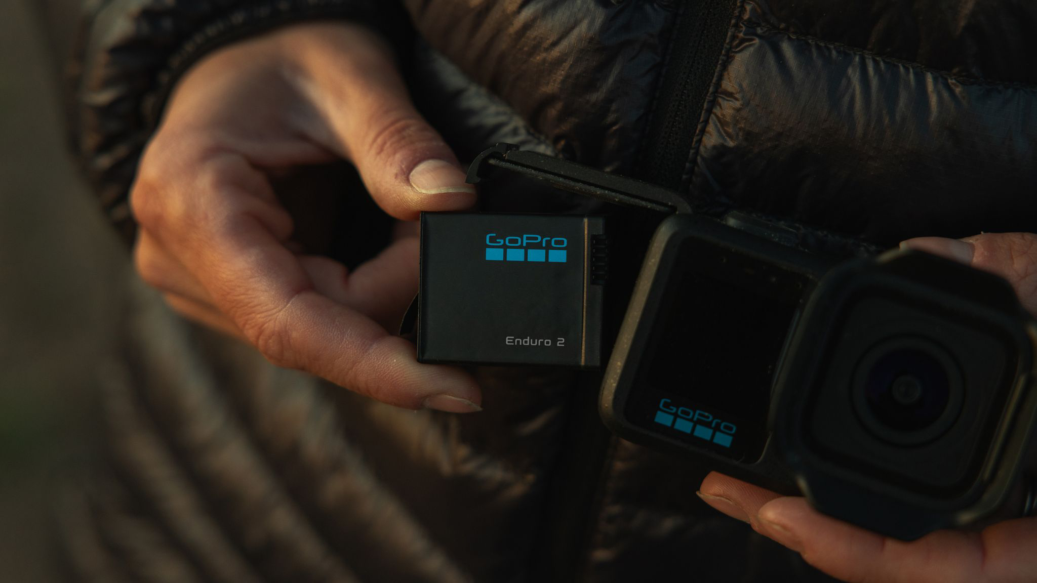 Egy kéz GoPro akkumulátort és kamerát tart. Fekete ruházat a háttérben.