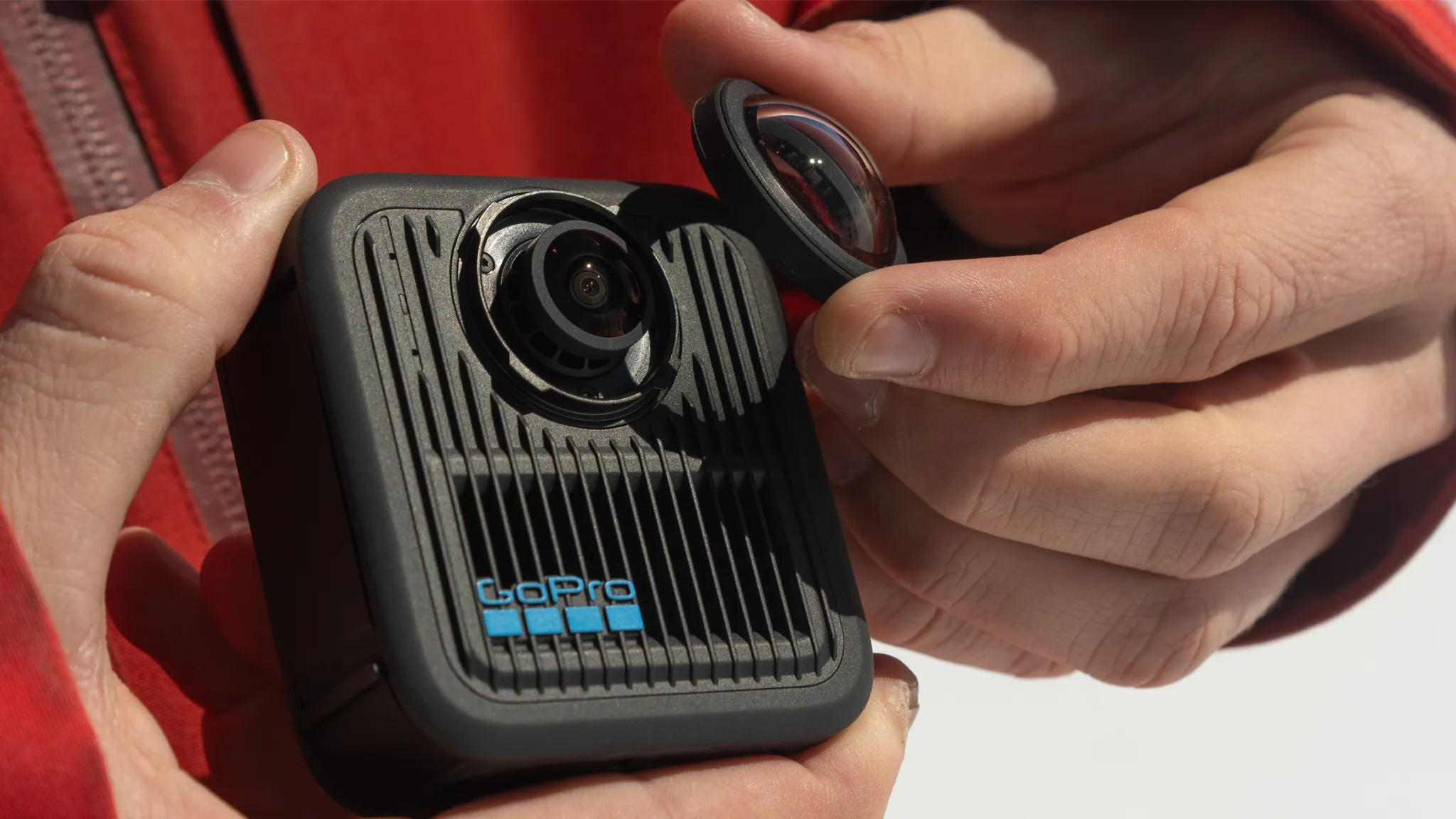 Kezek GoPro kamerát tartanak, eltávolított lencsével. Piros dzseki a háttérben.