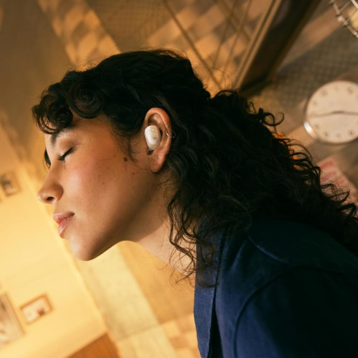 Una donna con gli occhi chiusi indossa un auricolare bianco. Un orologio è visibile sullo sfondo.