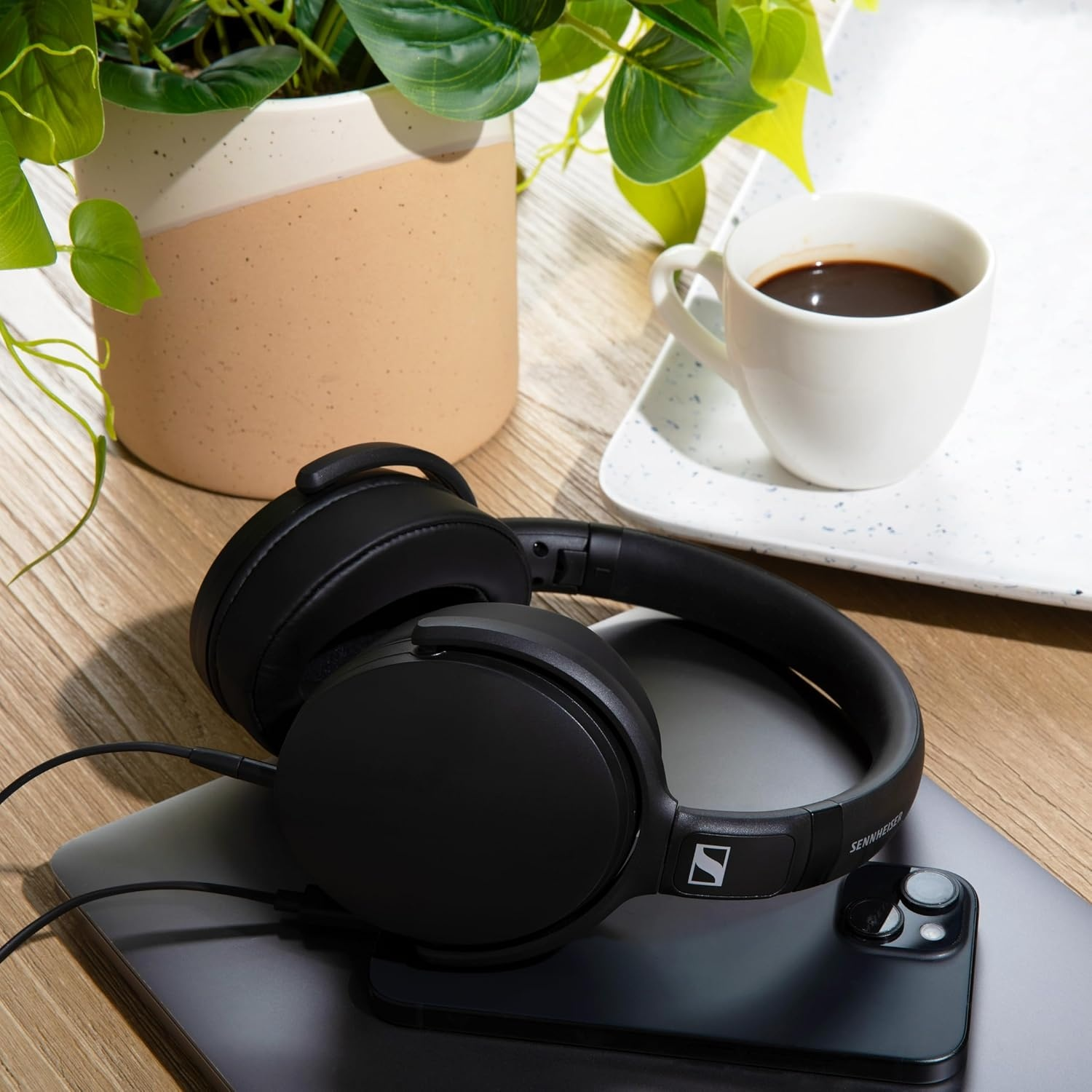 Cuffie nere su laptop e telefono, accanto a tazza di caffè e pianta.