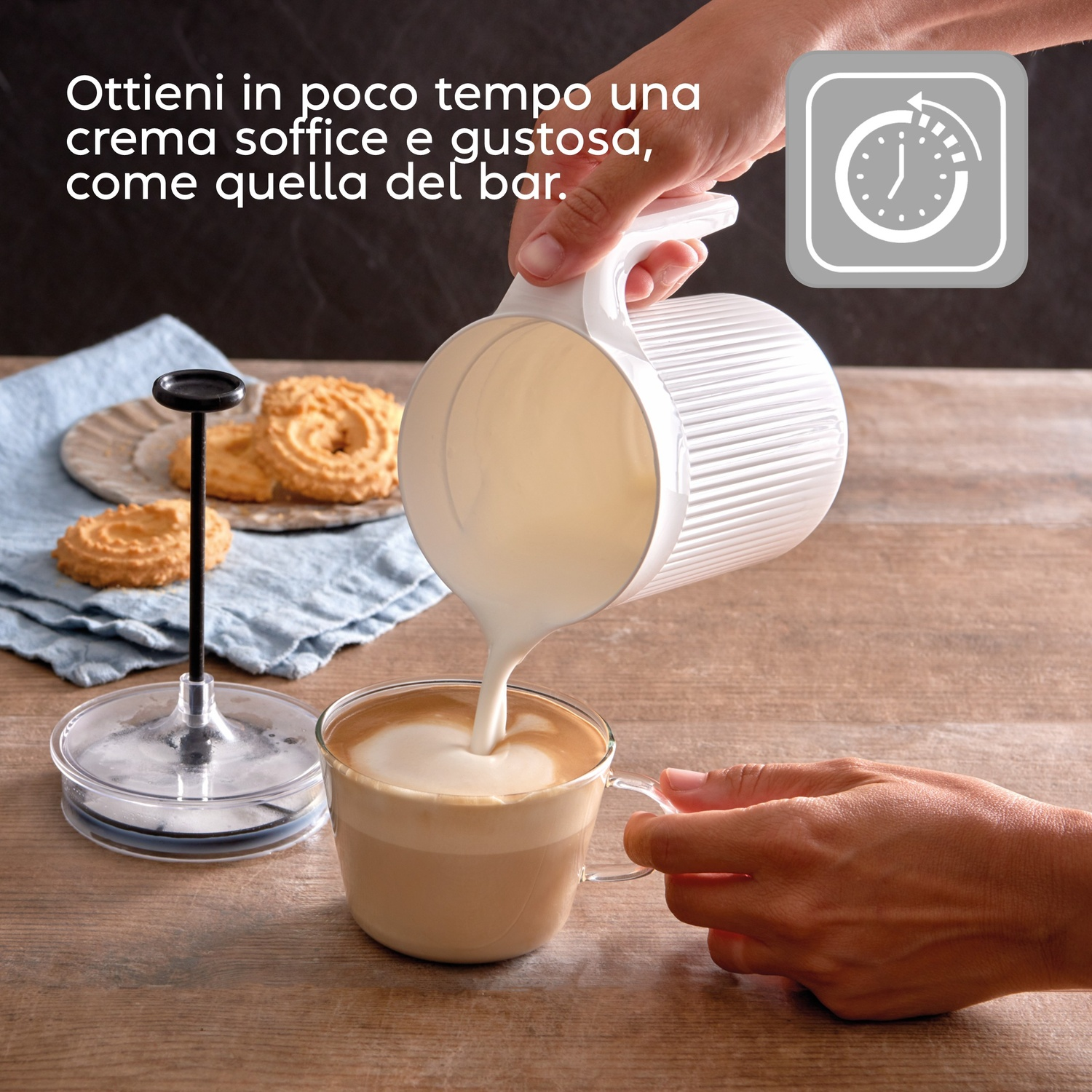 Una mano versa latte da un bricco bianco in una tazza di vetro con caffè latte art.