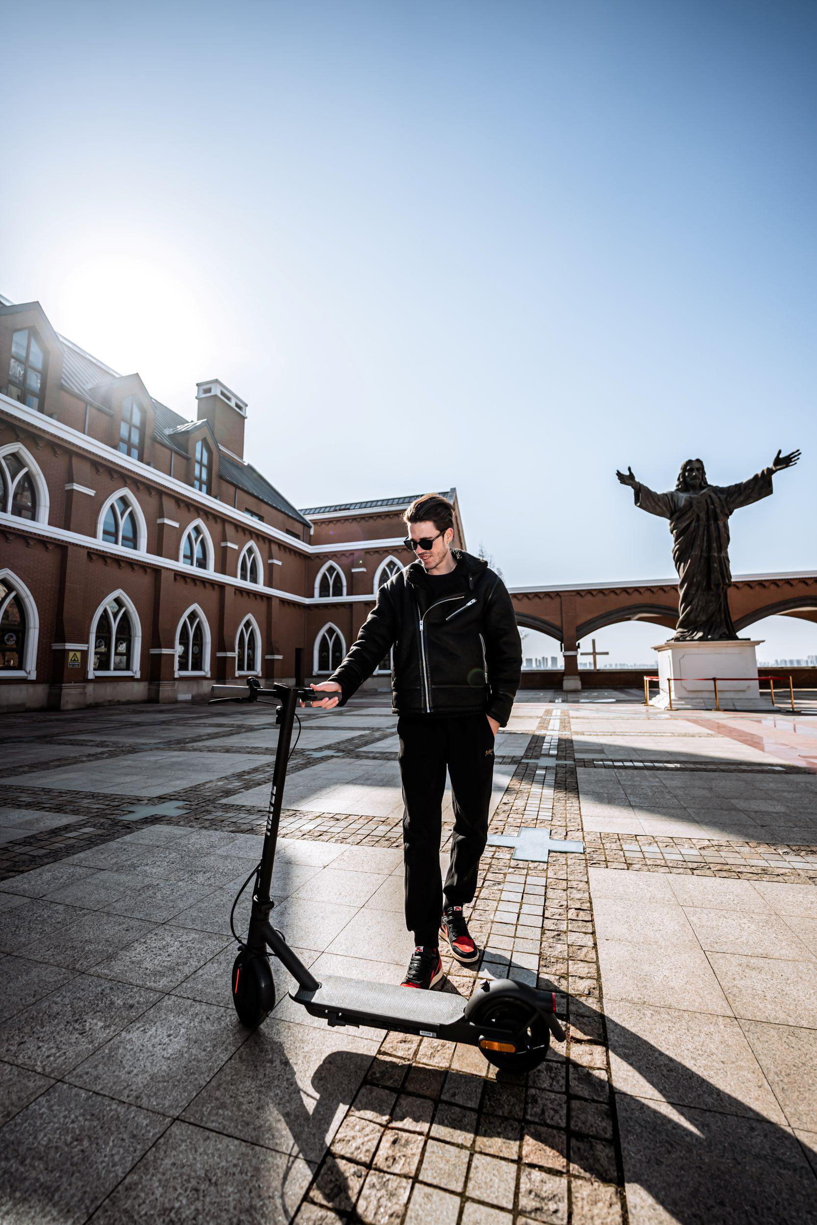 Ein Mann in schwarzer Kleidung steht neben einem E-Scooter vor einem Gebäude mit einer Christusstatue.