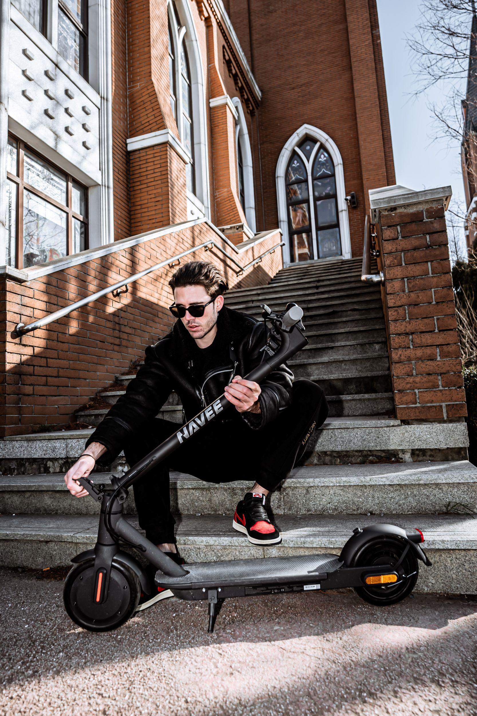Ein Mann in schwarzer Kleidung sitzt auf einer Steintreppe und hält einen schwarzen E-Scooter.