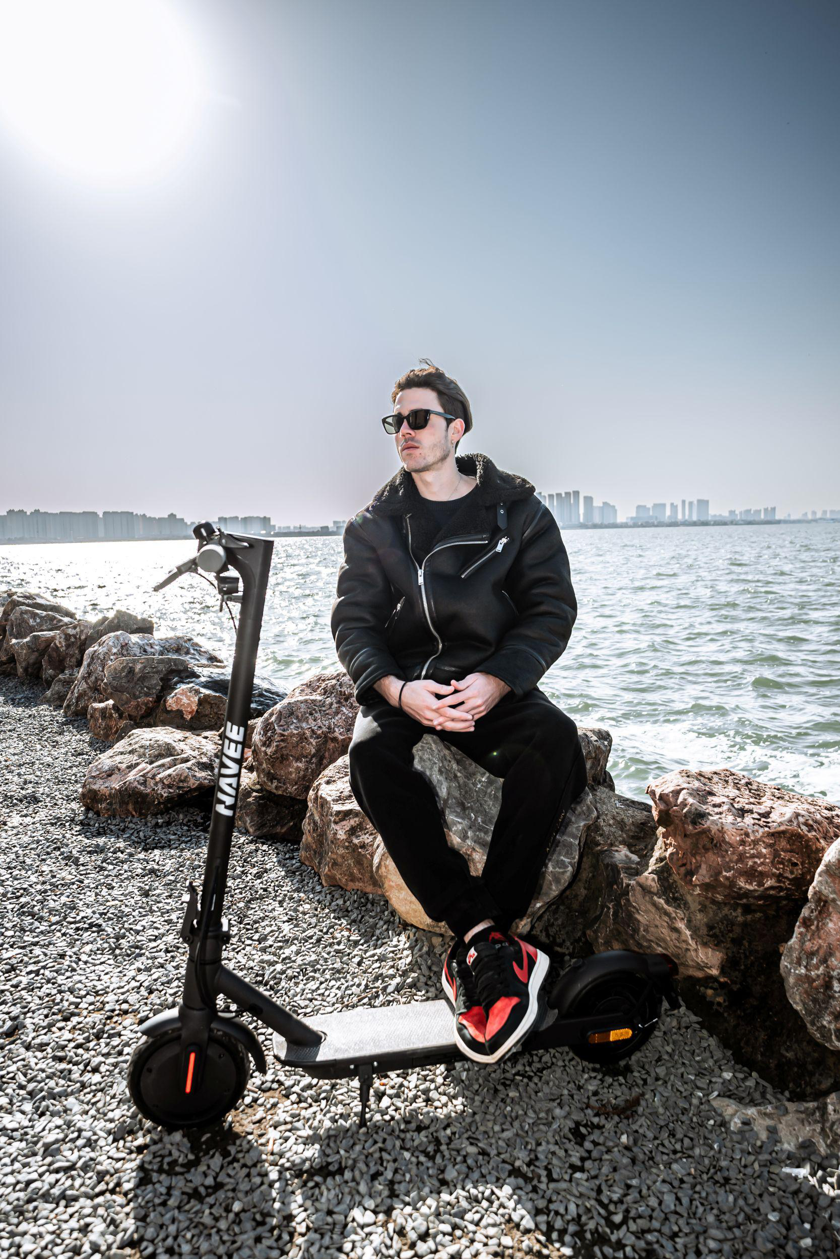 Ein Mann sitzt auf Felsen neben einem Roller, mit Blick aufs Wasser und eine Stadt.