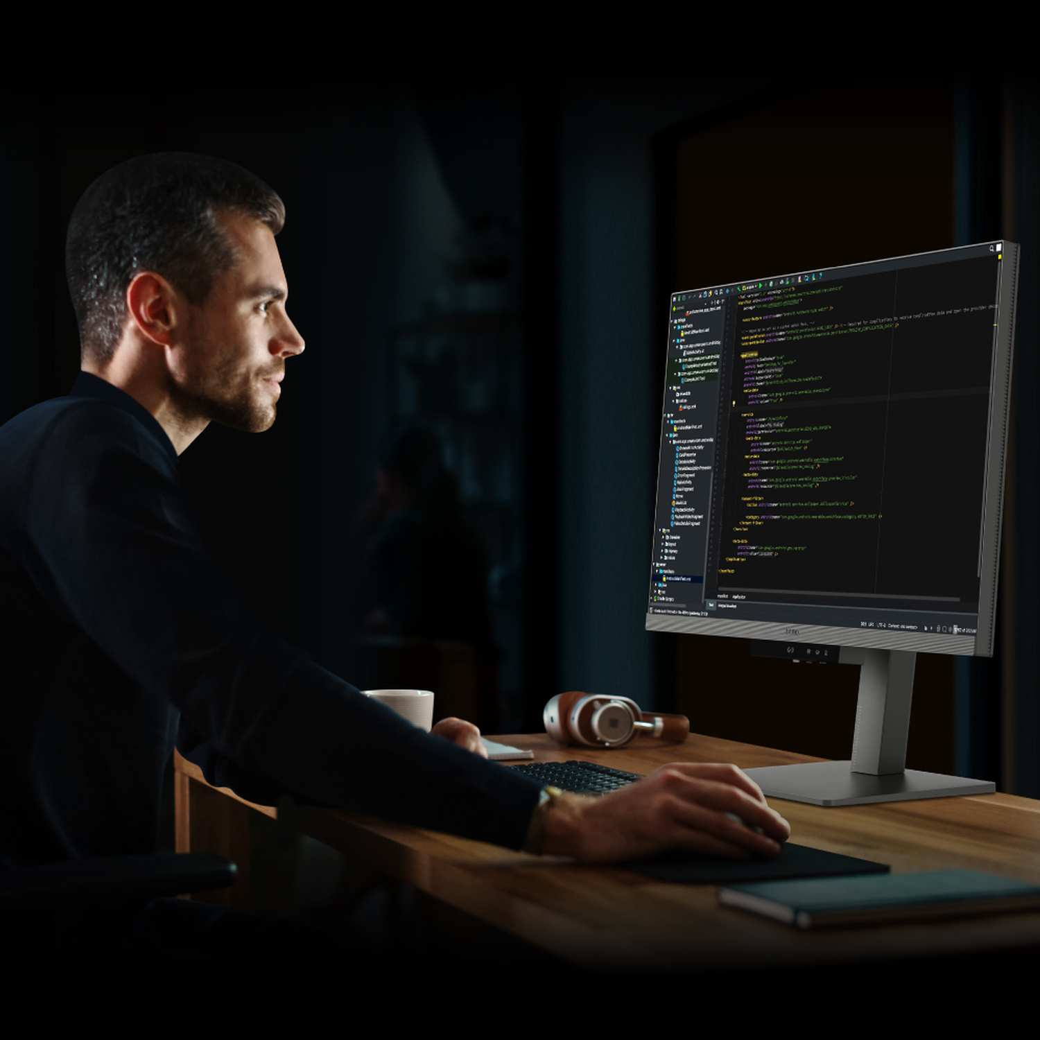 Un uomo siede a una scrivania, digitando codice su un monitor di computer. Cuffie vicine.