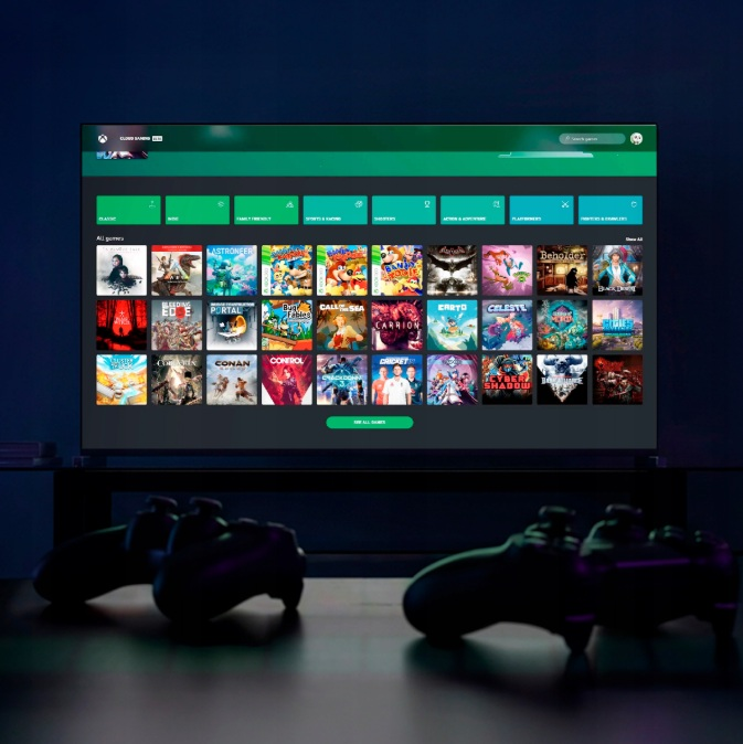 Ekran telewizora wyświetla menu gier Xbox z wieloma kafelkami gier. Dwa kontrolery są na pierwszym planie.