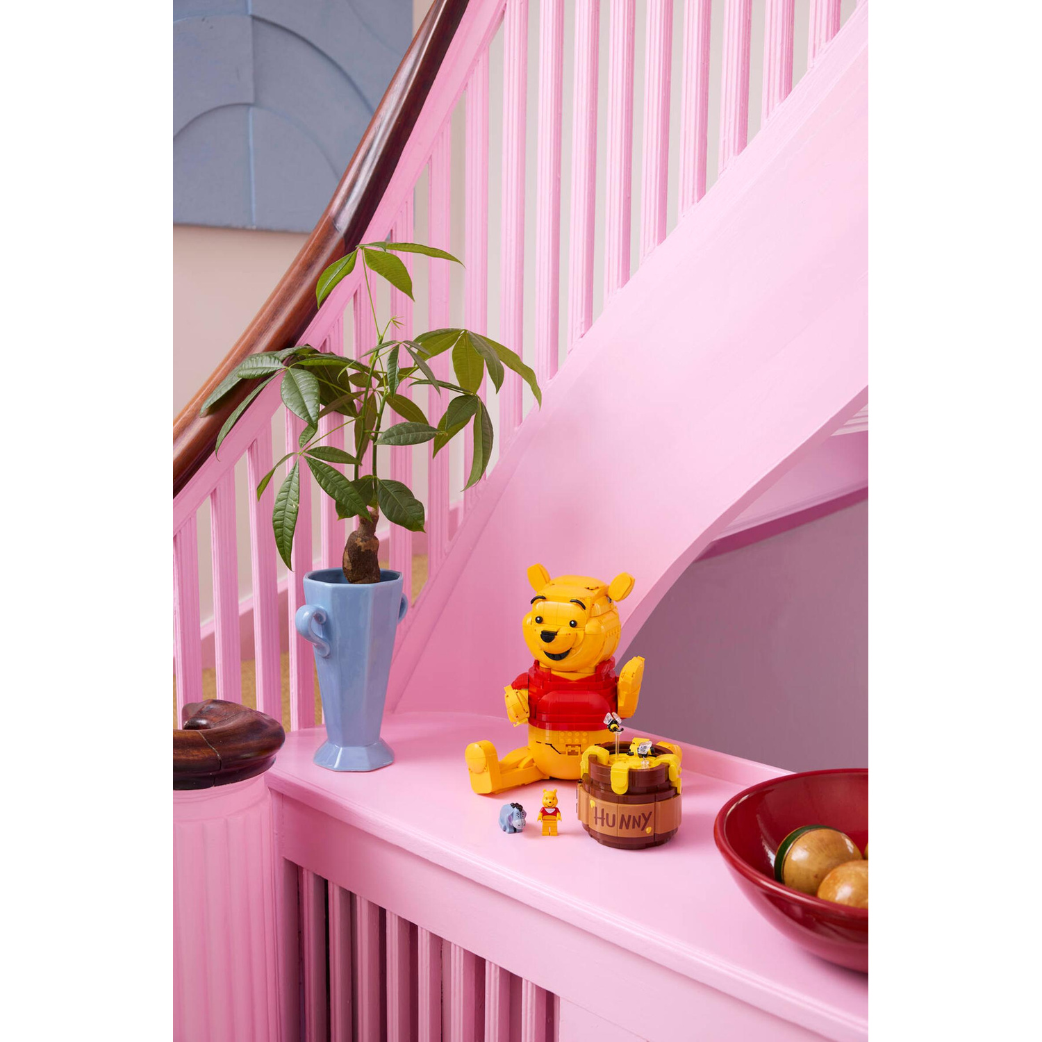 Statua di Winnie the Pooh con vaso di miele su scale rosa, vicino a pianta in vaso blu.