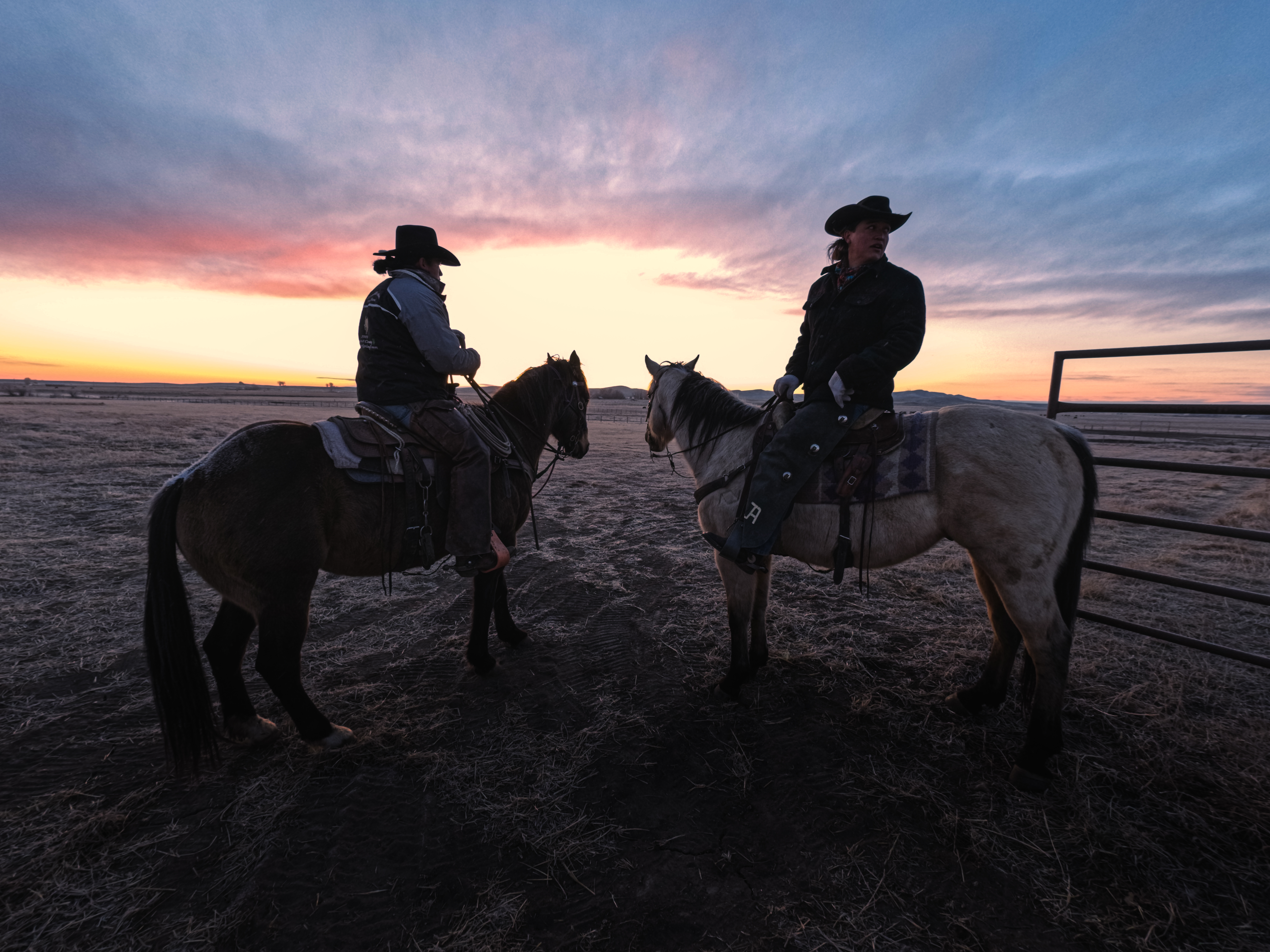 Zwei Cowboys auf Pferden bei Sonnenaufgang, mit buntem Himmel und Zaun.