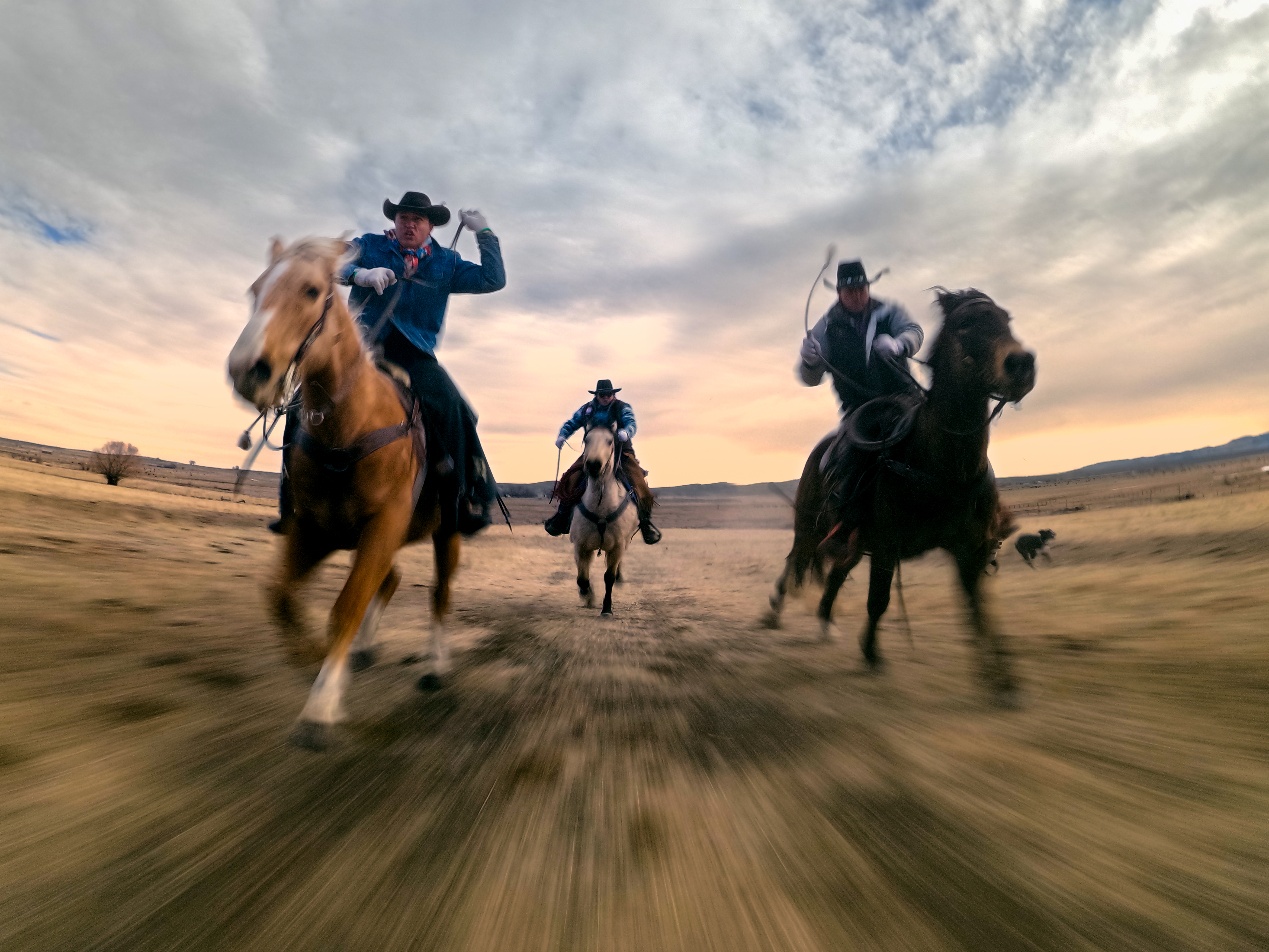 Drei Cowboys reiten auf Pferden im Galopp über eine staubige Ebene bei Sonnenuntergang.
