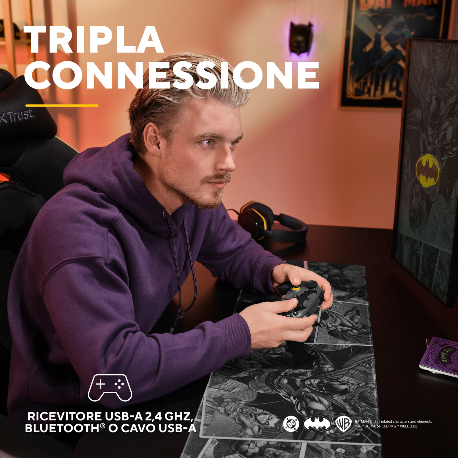 Uomo con felpa viola gioca con controller. Motivi di Batman sulla scrivania. Tripla connessione.