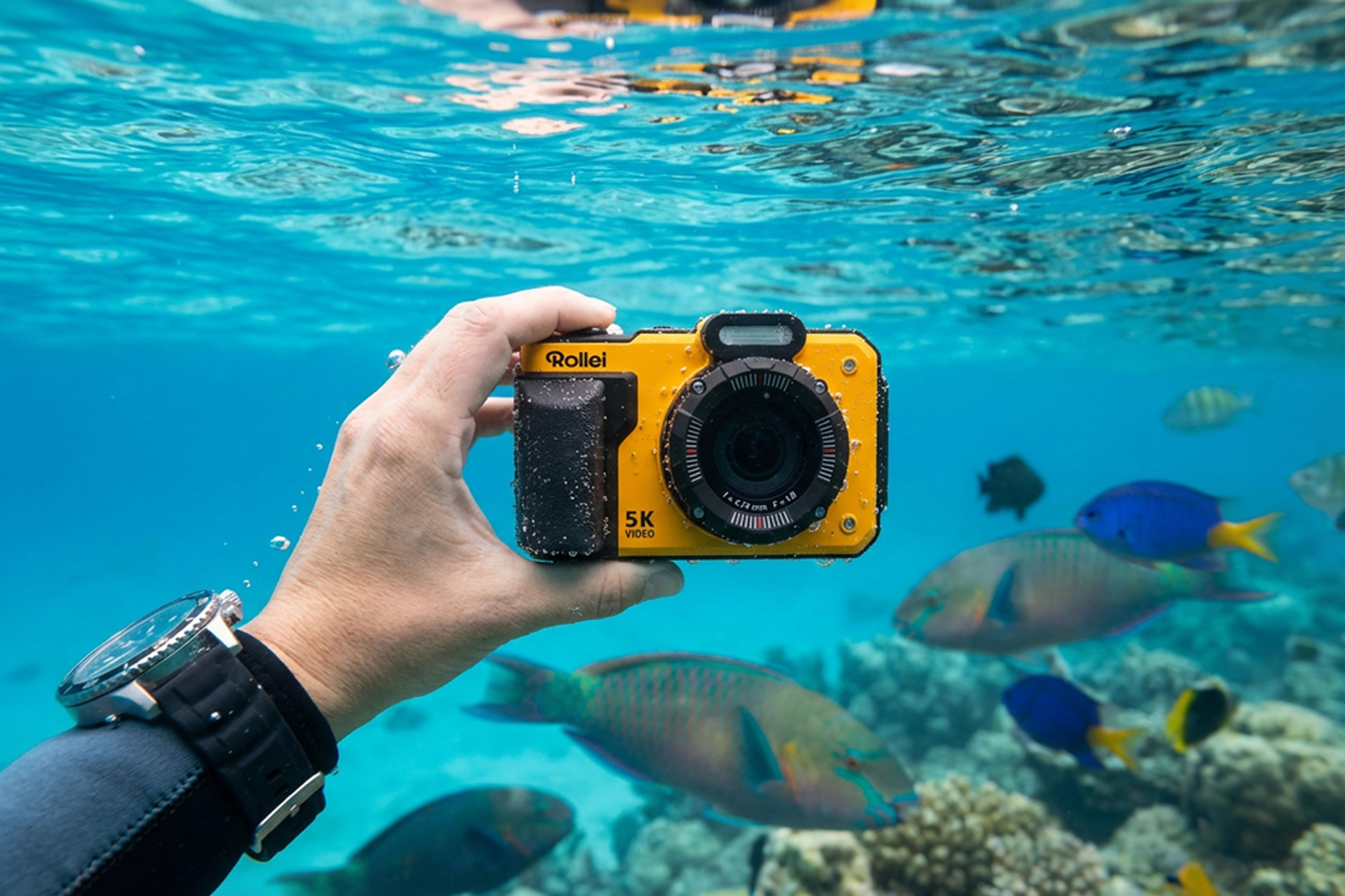 Gelbe Unterwasserkamera in Hand vor Korallenriff und Fischen.
