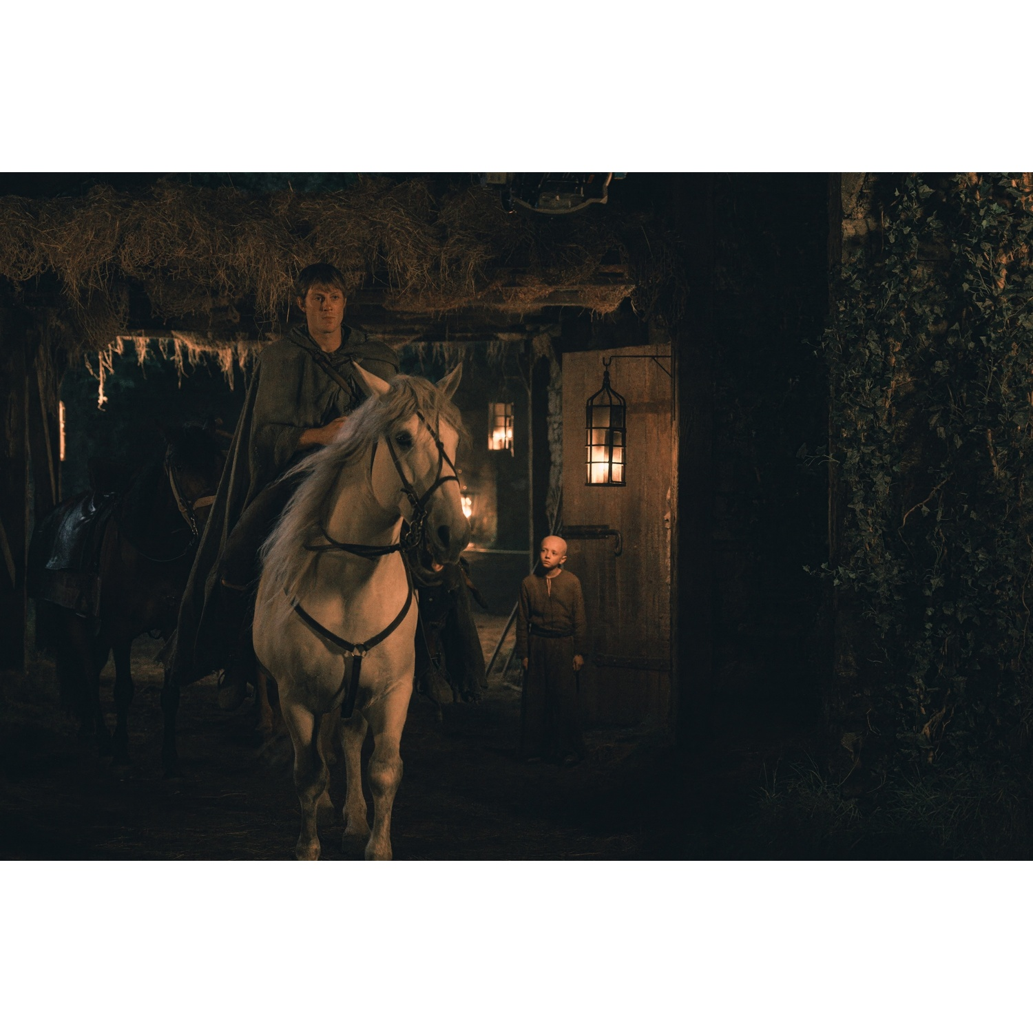 Homme en cape sur cheval blanc, garçon en tunique près de la porte de la grange.