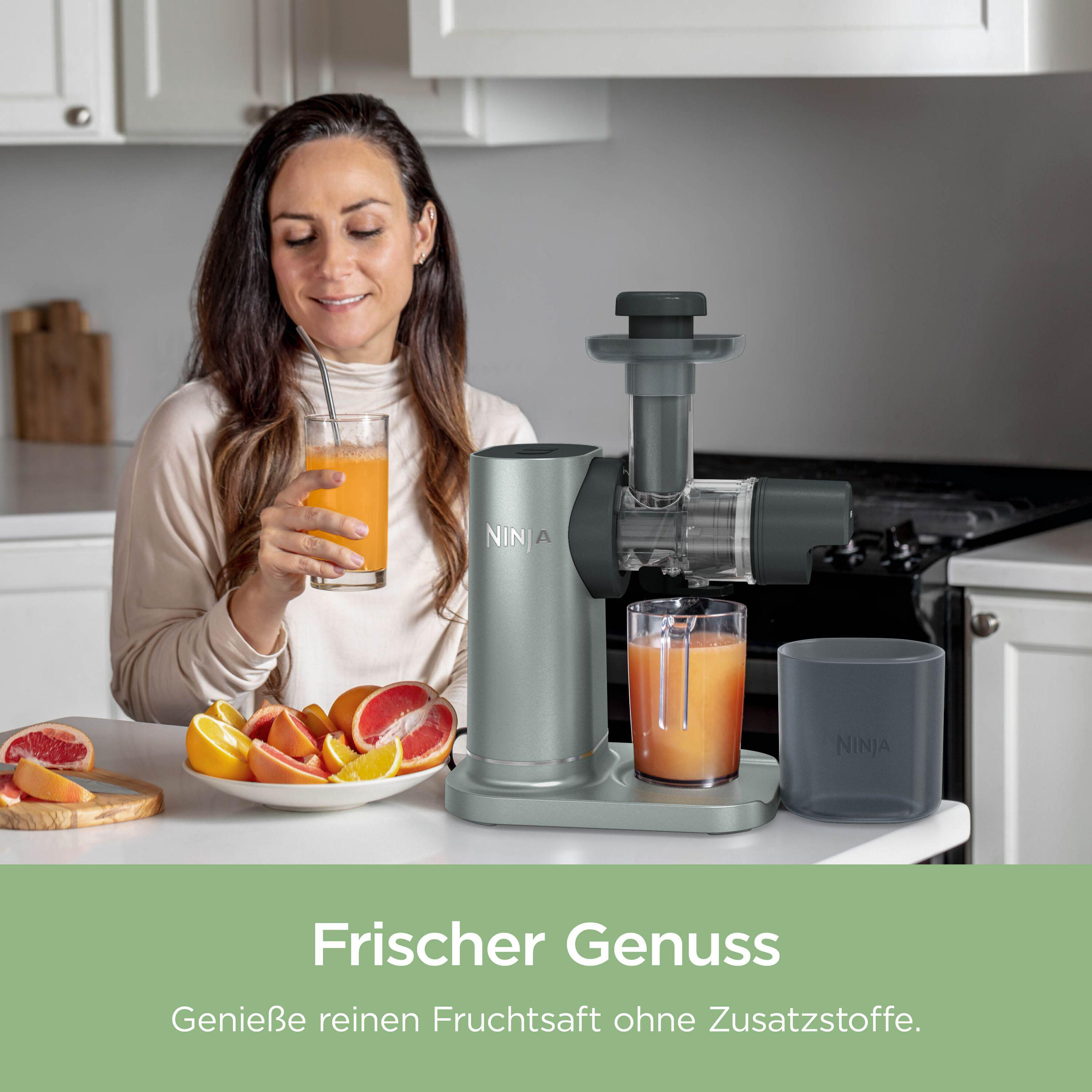 Eine Frau hält ein Glas Orangensaft neben einem Entsafter mit Zitrusfrüchten.