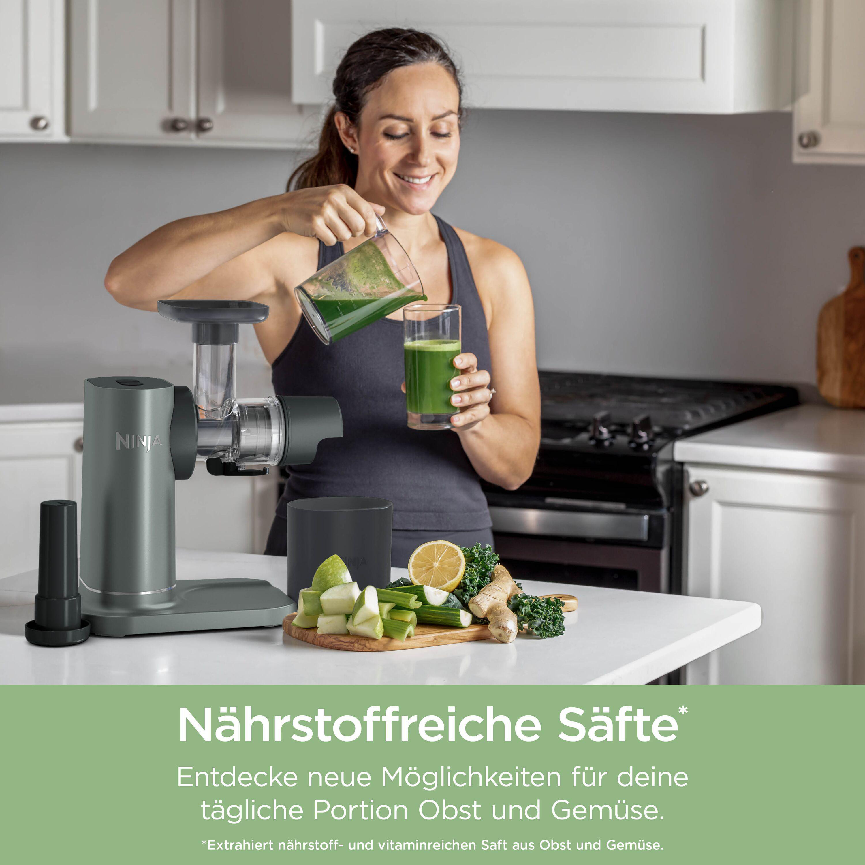 Eine Frau gießt grünen Saft in ein Glas, neben einem Entsafter und Zutaten.