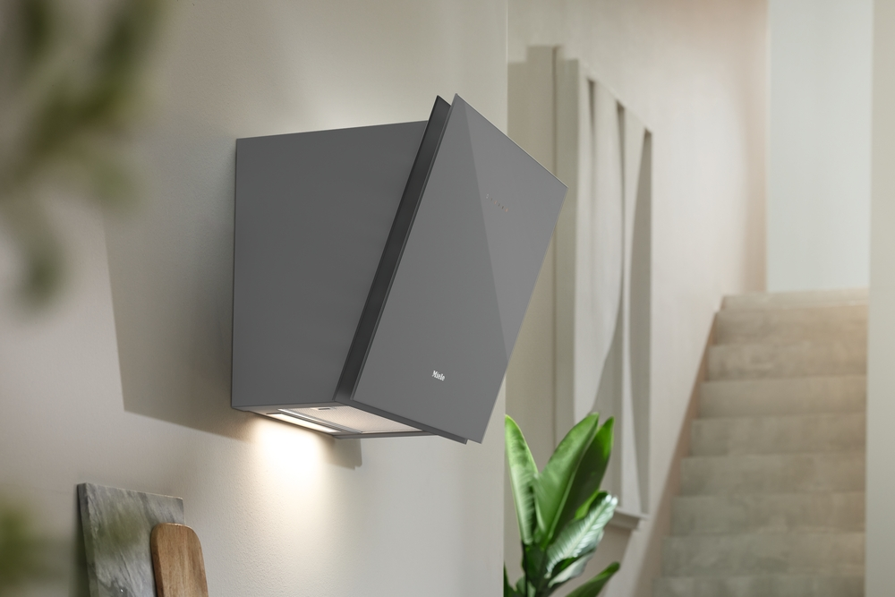 Grauer Miele Dunstabzugshaube an einer Wand, beleuchtet, mit Treppe und Pflanze.
