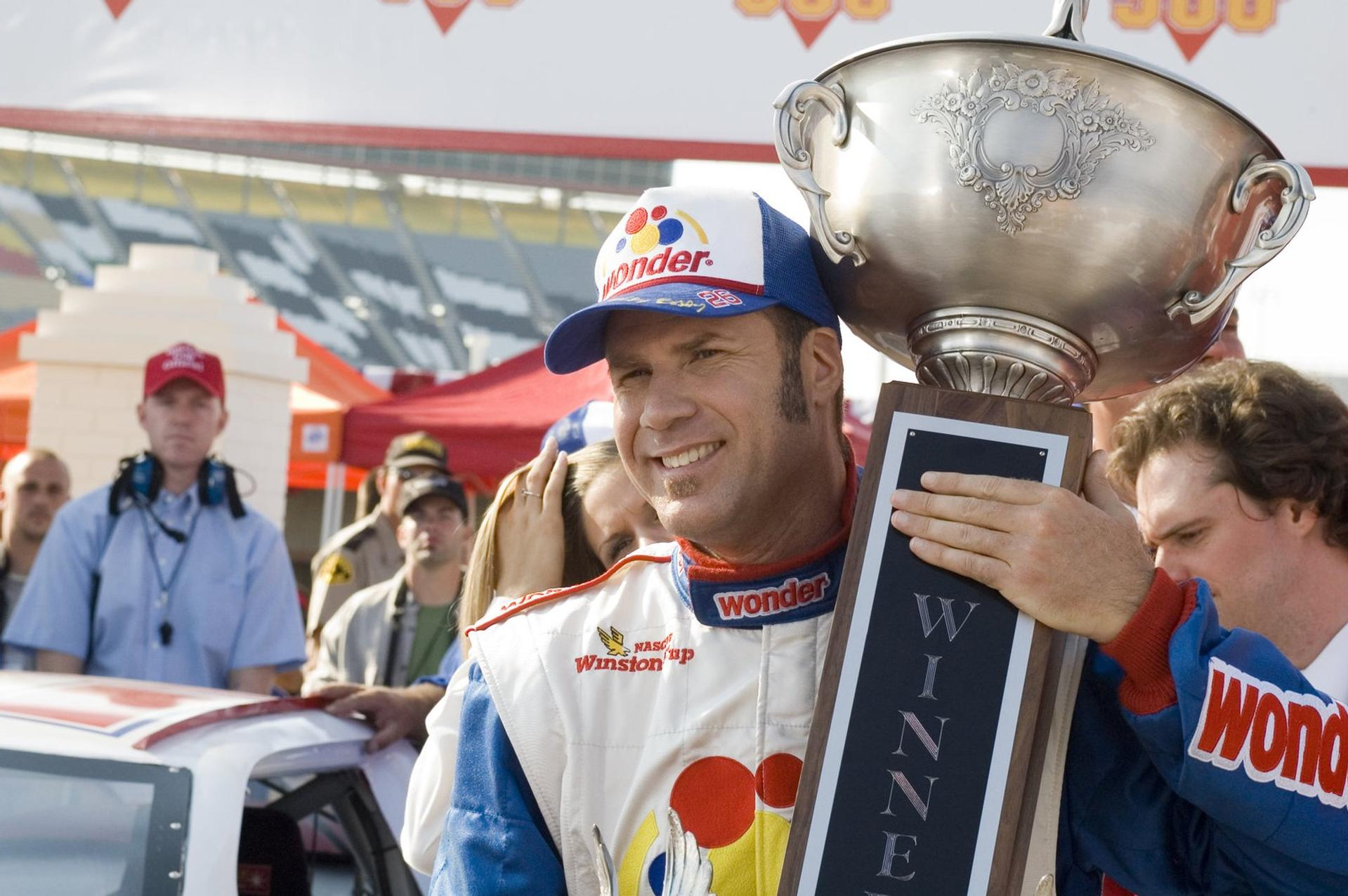 Ein Rennfahrer mit einem Pokal lächelt, hinter ihm sind Menschen und ein Stadion.