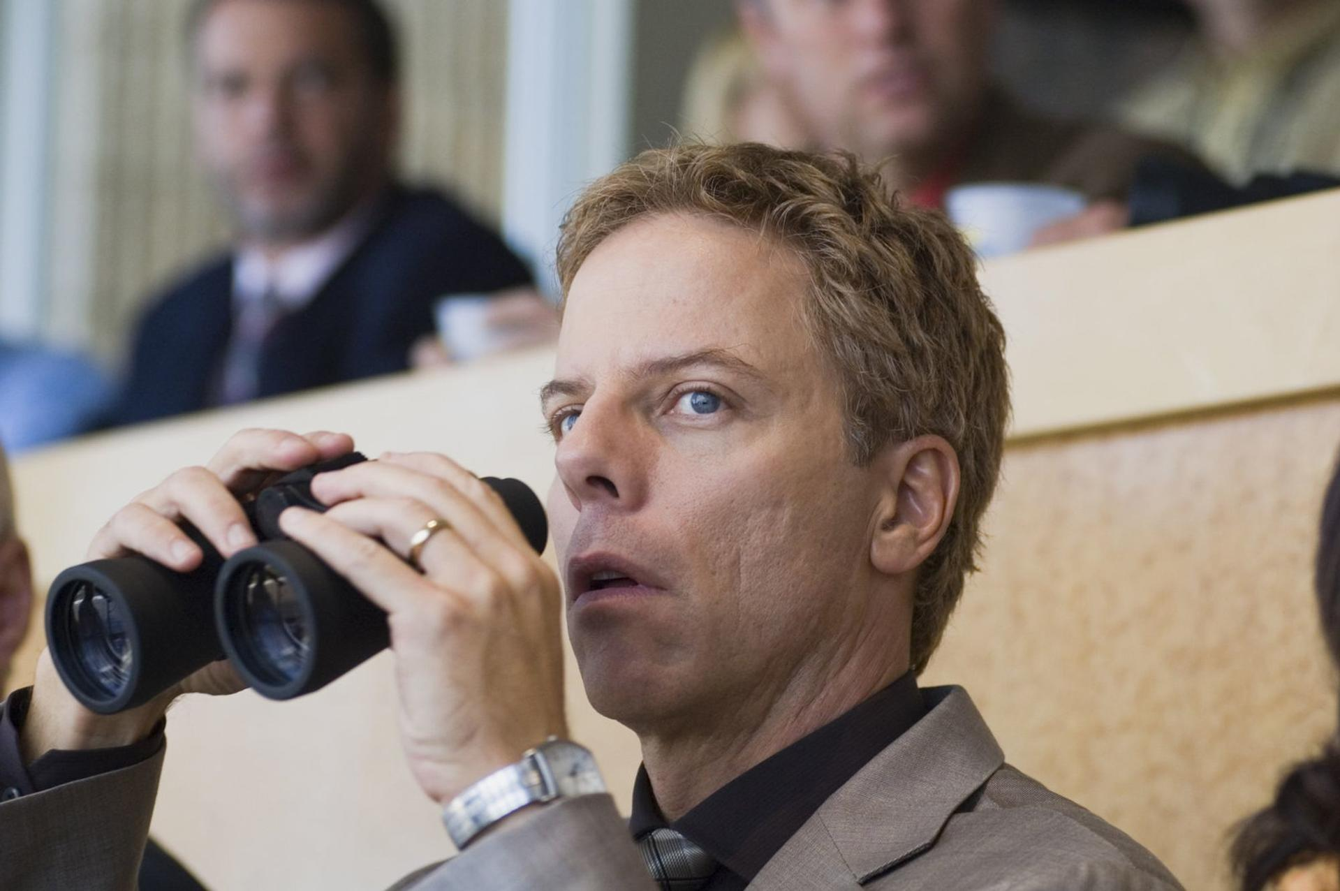 Ein Mann mit blonden Haaren schaut durch ein Fernglas. Im Hintergrund unscharfe Personen.