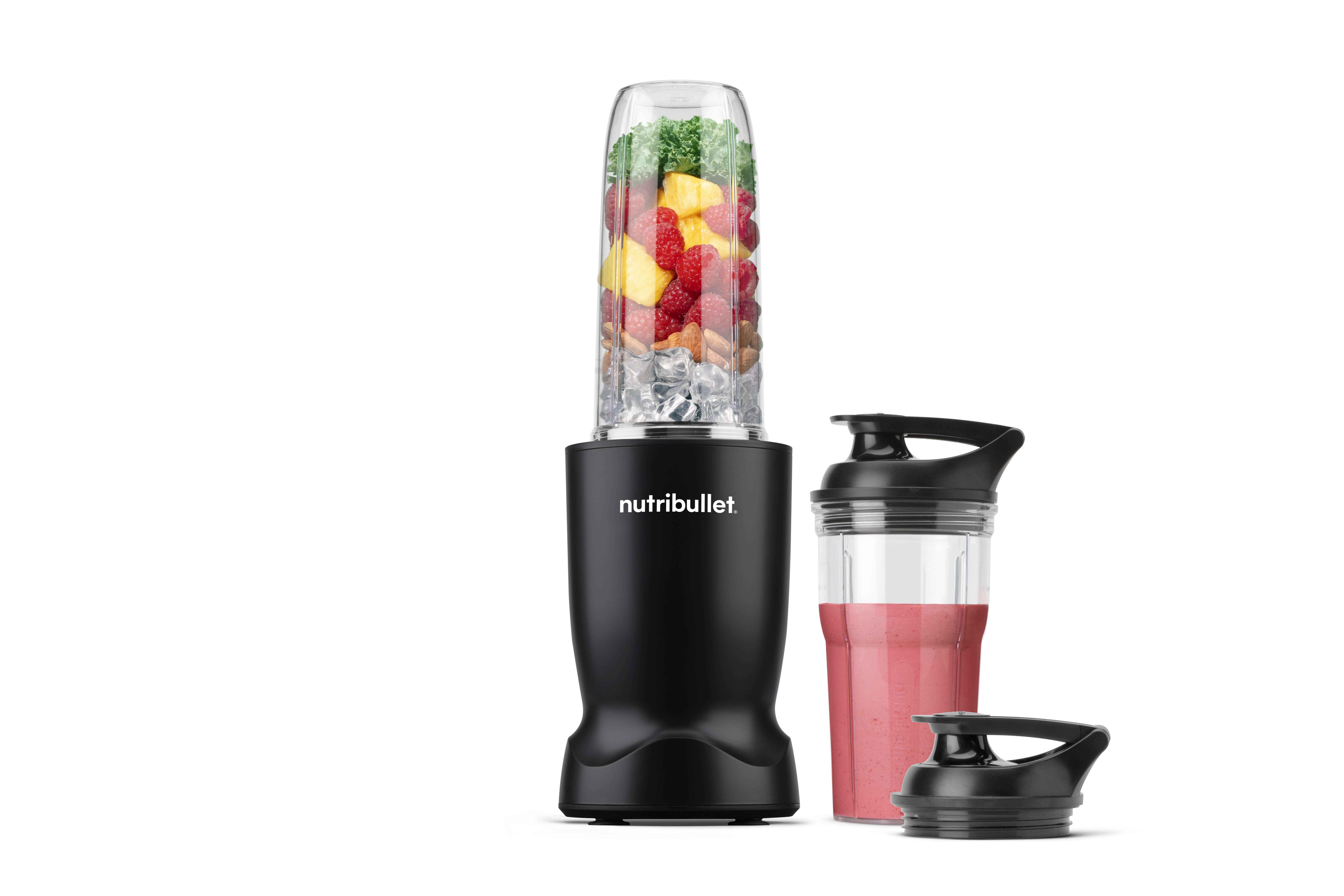 Egy fekete Nutribullet turmixgép jéggel, bogyókkal, ananásszal és kelkáposztával egy pohárban, egy turmixos pohár mellett.